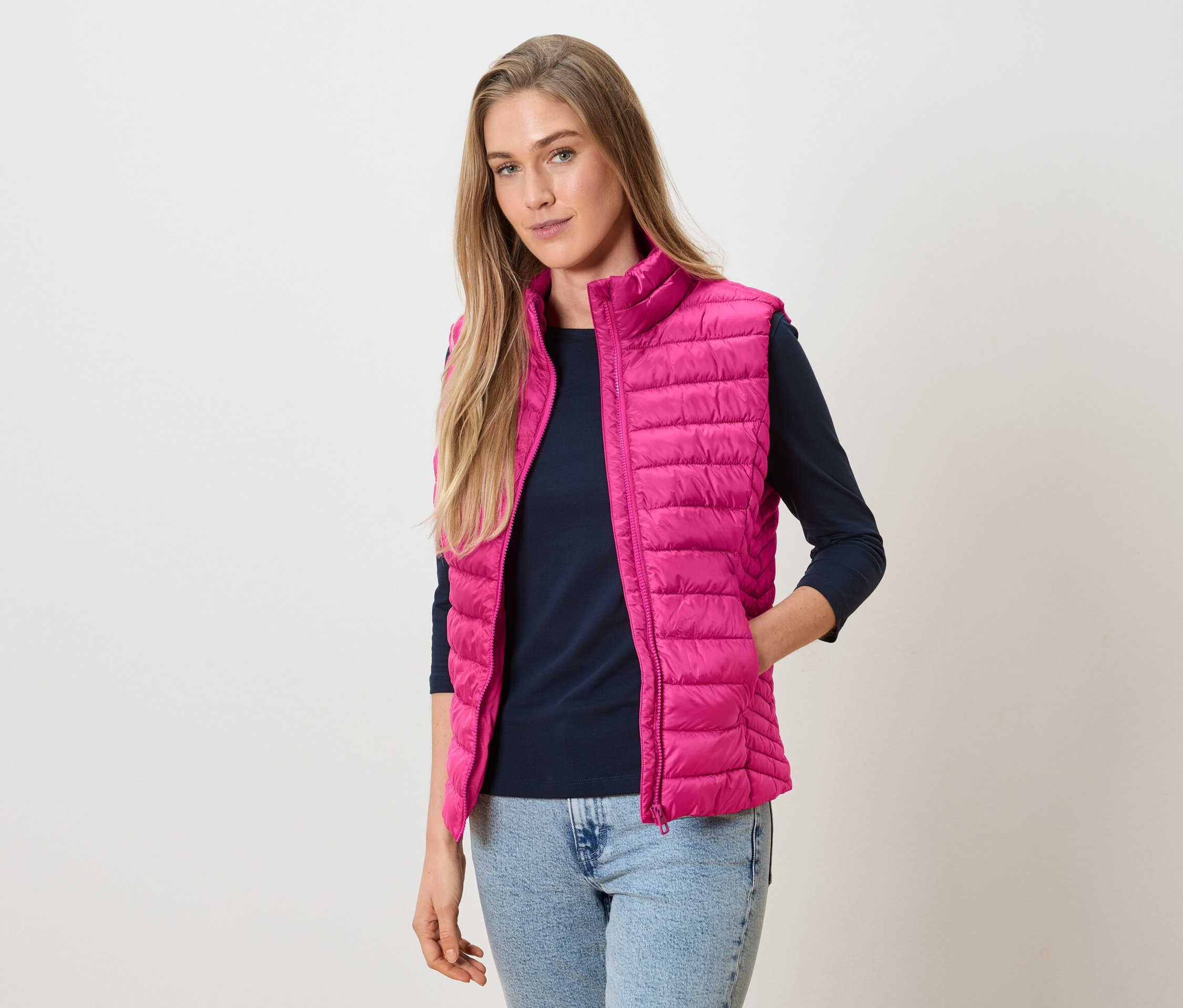 Žena má na sebe ružovú prešívanú vestu cez tmavomodré tričko s dlhým rukávom a džínsy.