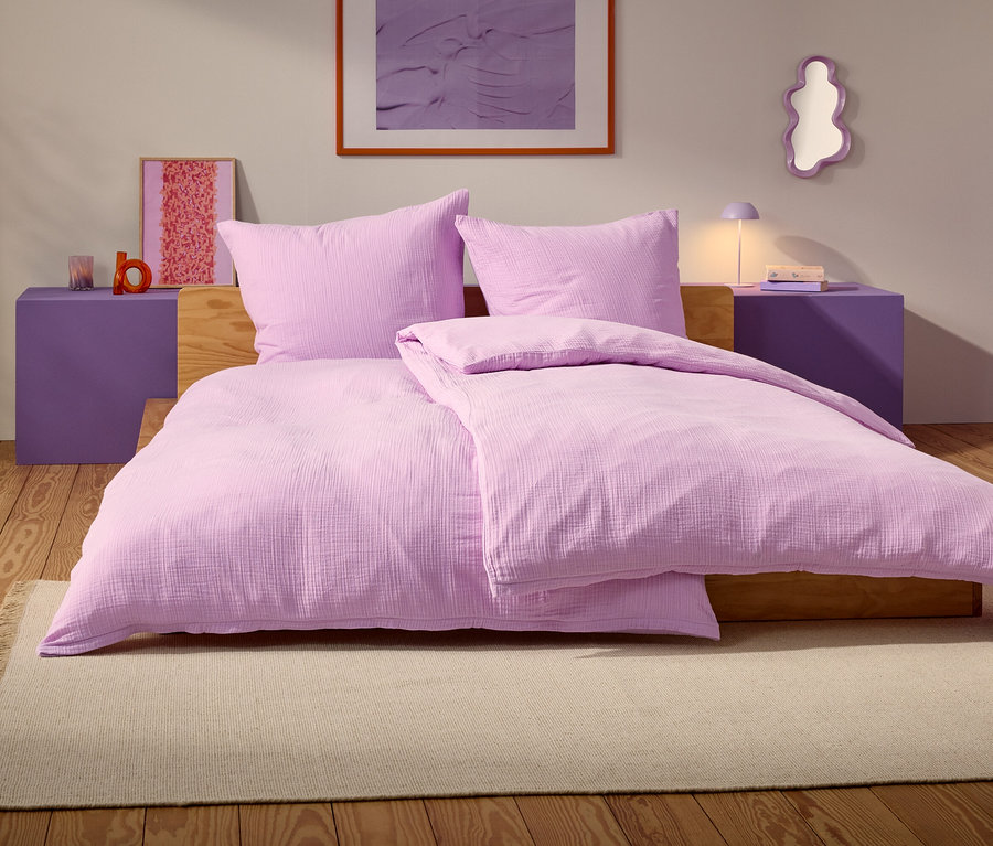 Fialová posteľ s dvoma vankúšmi a prikrývkou stojí na béžovom koberci v spálni.