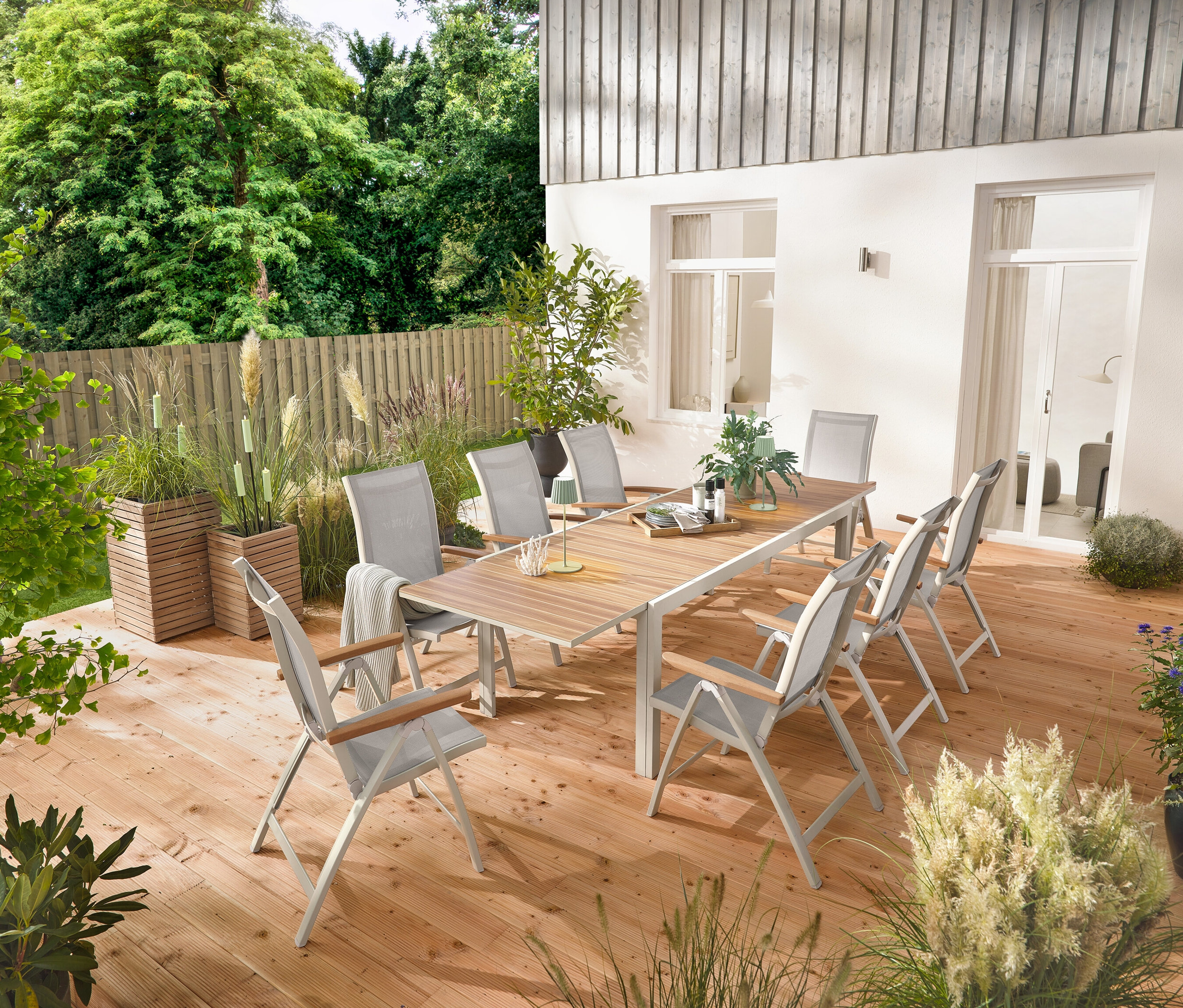 Rozkladací hliníkový záhradný stôl »Liska«, cca 200 – 300 cm dlhý, stojí na drevenej terase, obklopený šiestimi vysokými kreslami »Liska« s nastaviteľným operadlom. Viditeľné sú aj 2 stĺpiky na rastliny »Tinus« a 5 LED záhradných fakieľ.