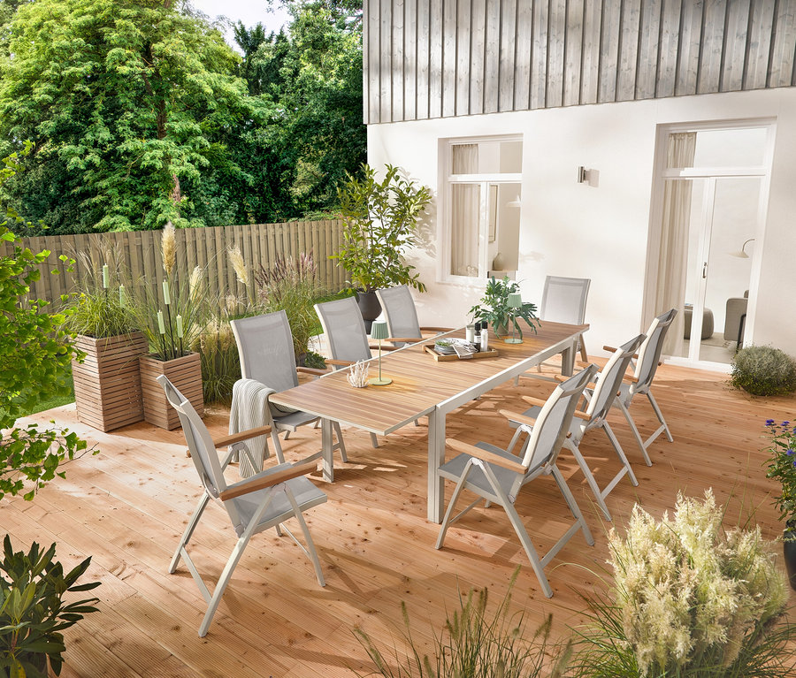 Rozkladací hliníkový záhradný stôl »Liska«, cca 200 – 300 cm dlhý, stojí na drevenej terase, obklopený šiestimi vysokými kreslami »Liska« s nastaviteľným operadlom. Viditeľné sú aj 2 stĺpiky na rastliny »Tinus« a 5 LED záhradných fakieľ.