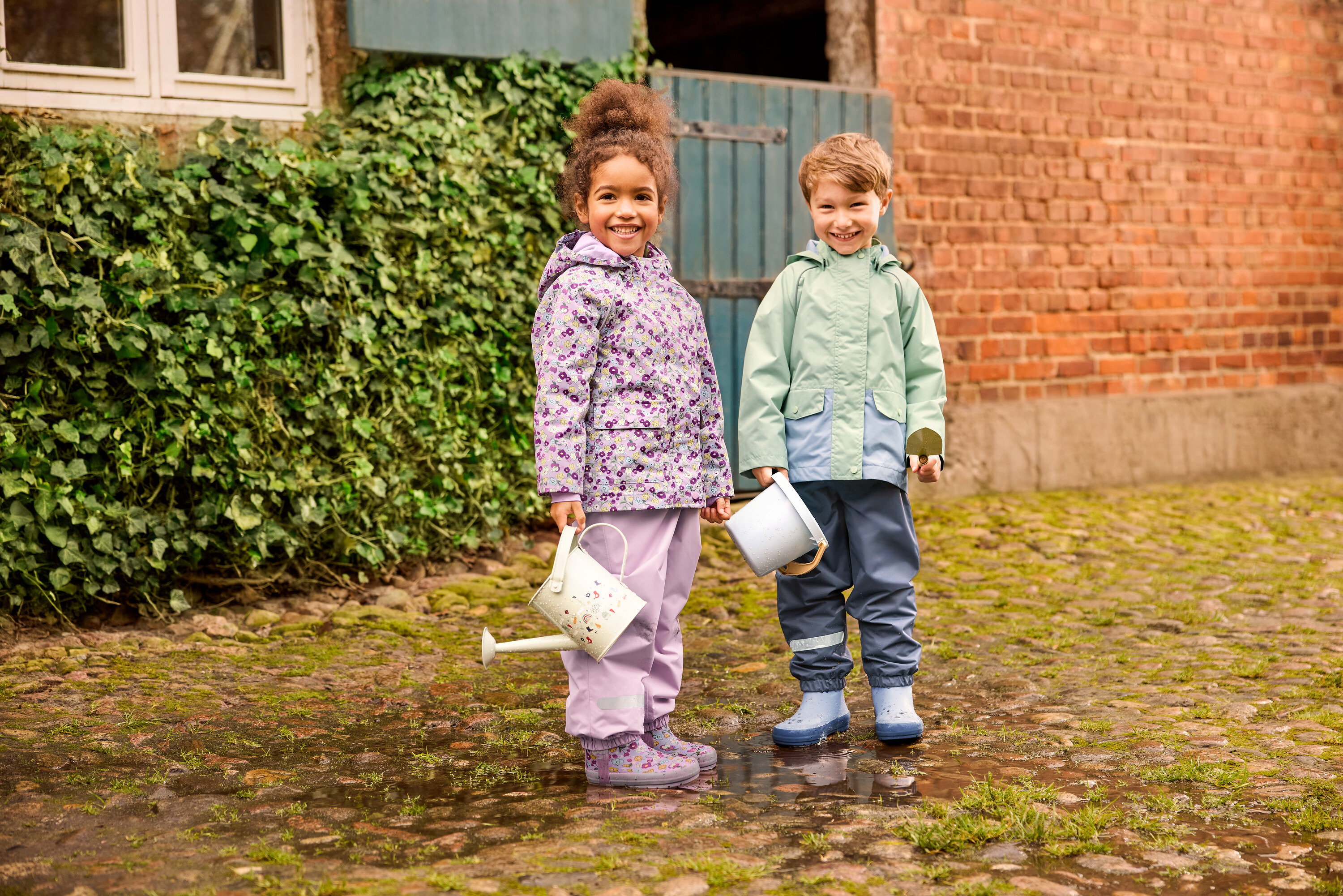 Dve deti stoja vonku: dievča v kvetinovej detskej dažďovej bunde a kvetinových detských gumákoch a chlapec v zelenej detskej dažďovej bunde a modrých detských gumákoch. Oboje majú na sebe detské dažďové nohavice.