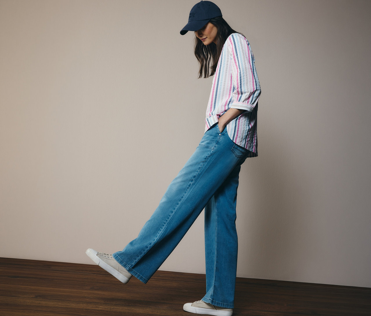 Žena v džínsoch a šiltovke má na sebe košeľovú blúzku CECIL s pruhmi. Stojí na drevenej podlahe.