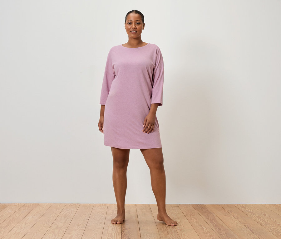 Žena stojí bosá pred stenou a má na sebe ružové šaty s krátkym rukávom.