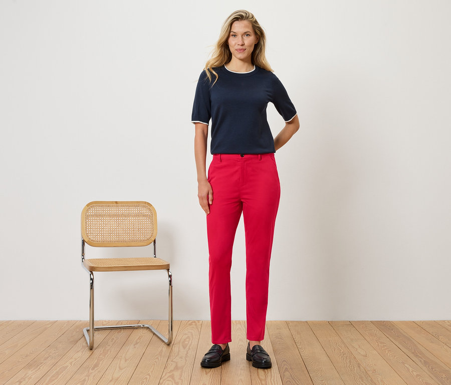 Žena stojí pred stenou vedľa stoličky. Má na sebe tmavomodrú blúzku s bielymi detailmi a červené nohavice.