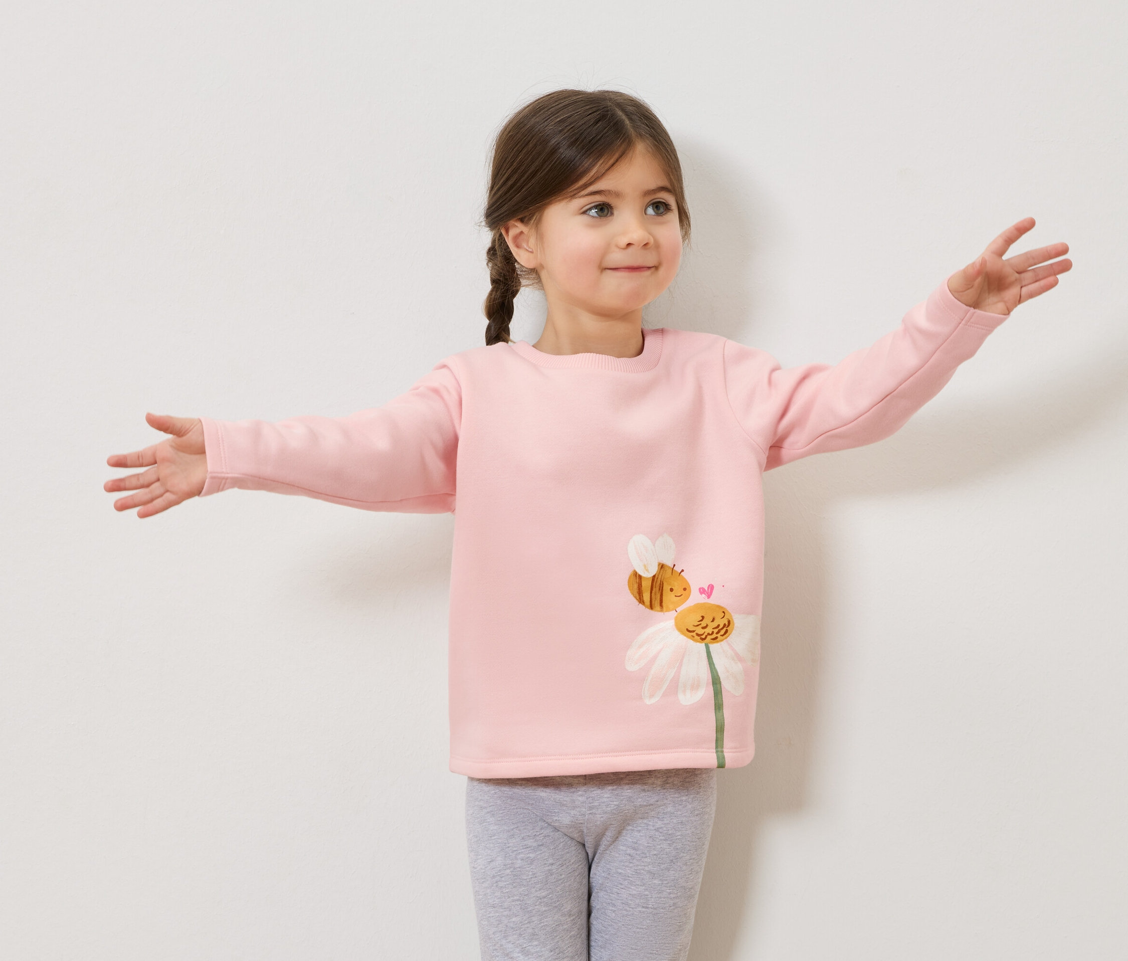 Dievča so zapletenými vlasmi v ružovom svetri s potlačou včiel a margarétok, sivé legíny, s vystretými rukami.