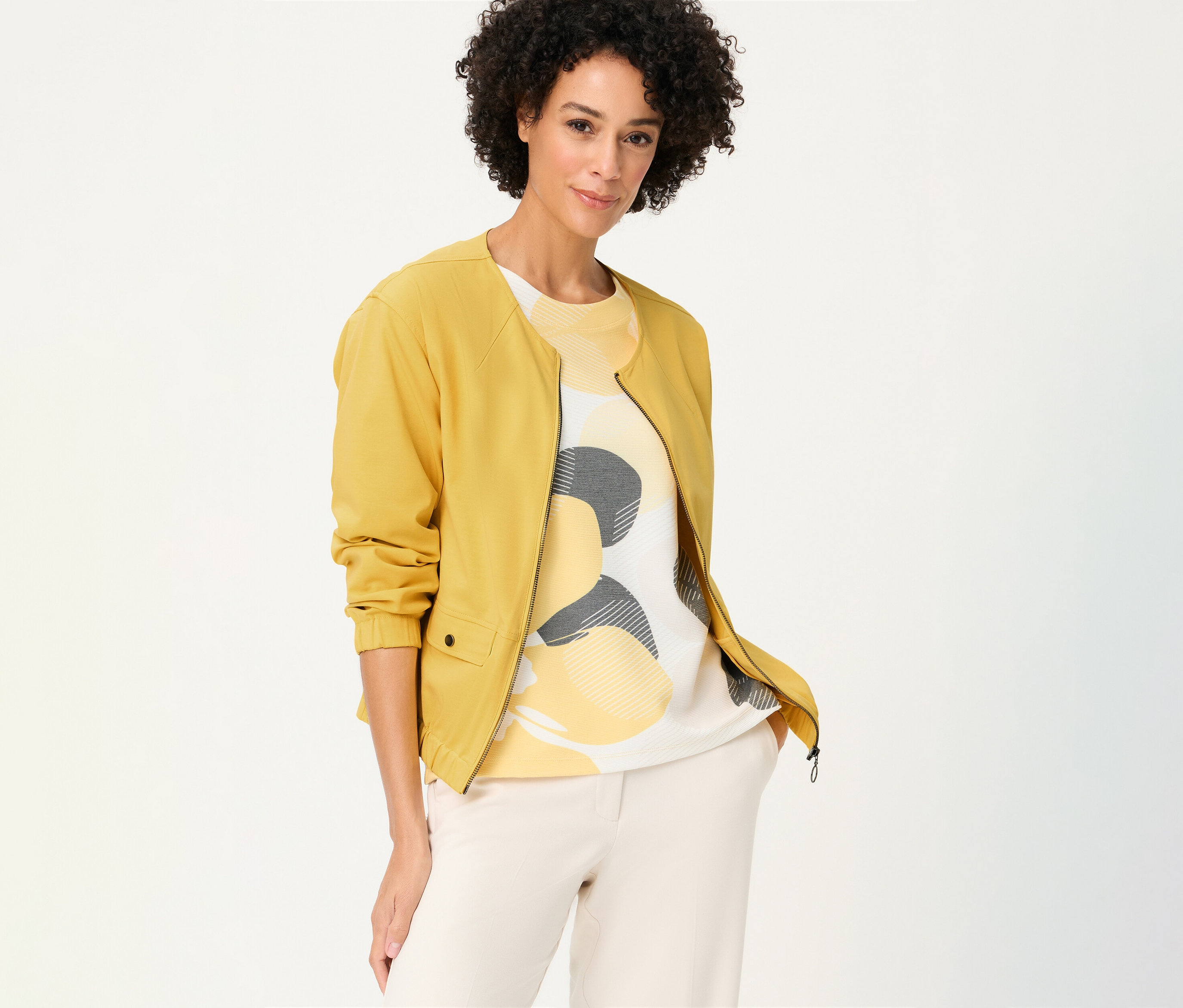 Žena s kučeravými vlasmi má na sebe žltú teplákovú bundu Olsen cez vzorovaný top a svetlé nohavice.