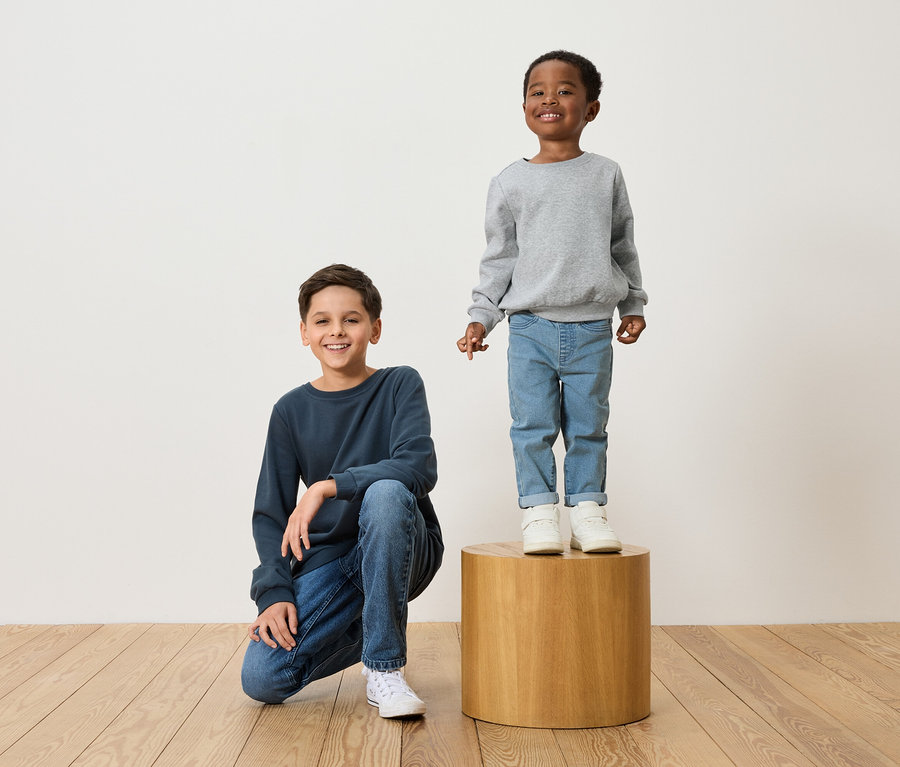 Dvaja chlapci pózujú: jeden kľačí v tmavomodrej mikine a džínsoch, druhý stojí na drevenej stoličke v šedej mikine a džínsoch.