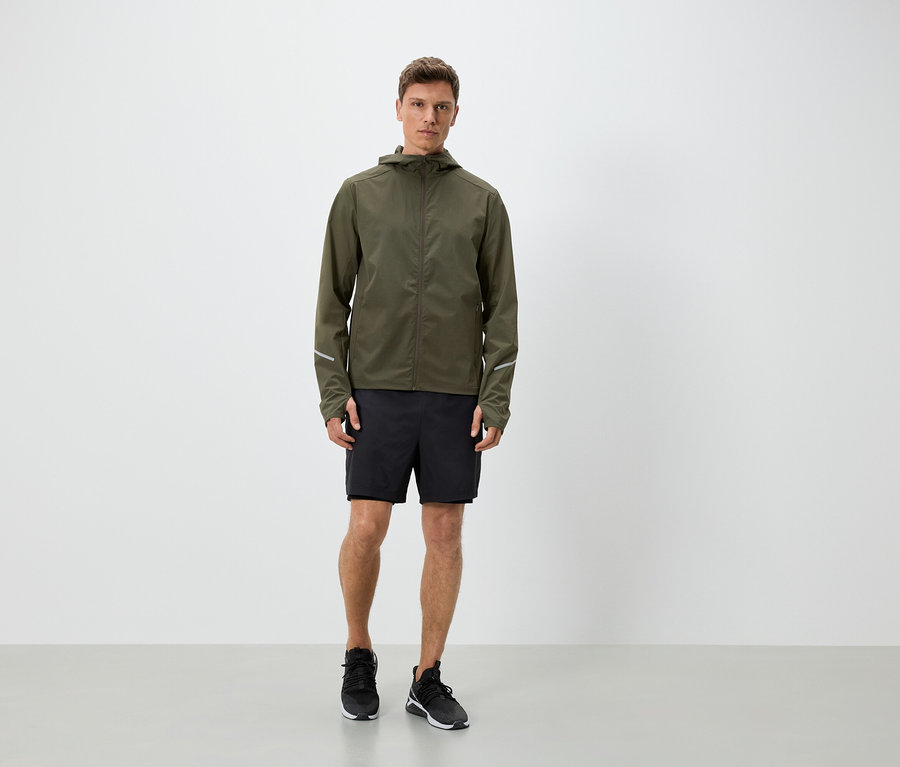 Muž v olivovozelenej bunde s kapucňou, čiernych šortkách a teniskách.