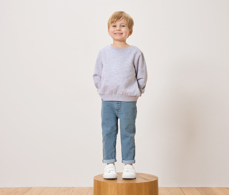 Usmievavý chlapec s blond vlasmi stojí na drevenej stoličke v sivom svetri, džínsoch a bielych teniskách.