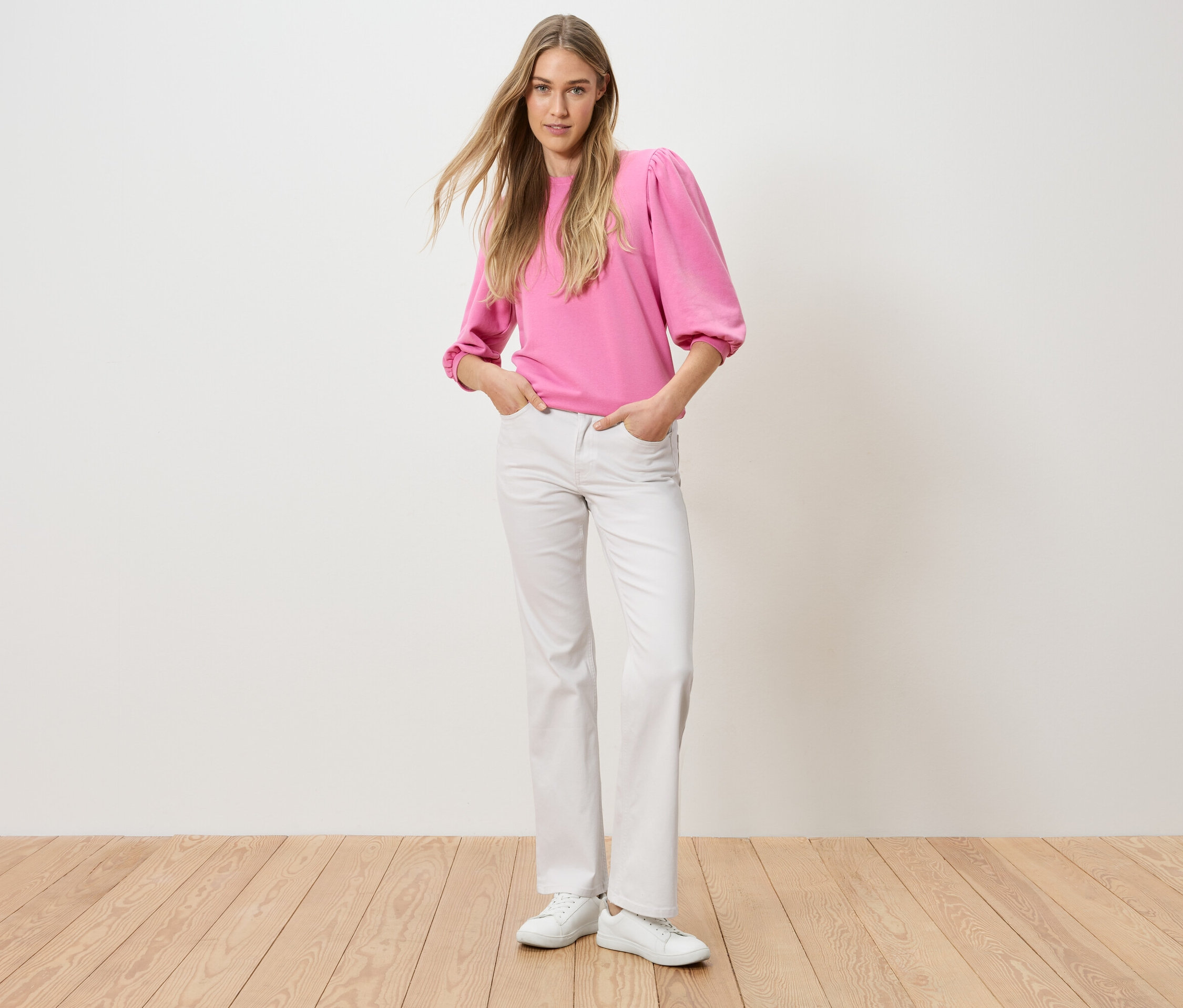 Žena stojí s rukami vo vreckách nohavíc. Má na sebe ružový top a biele džínsy.