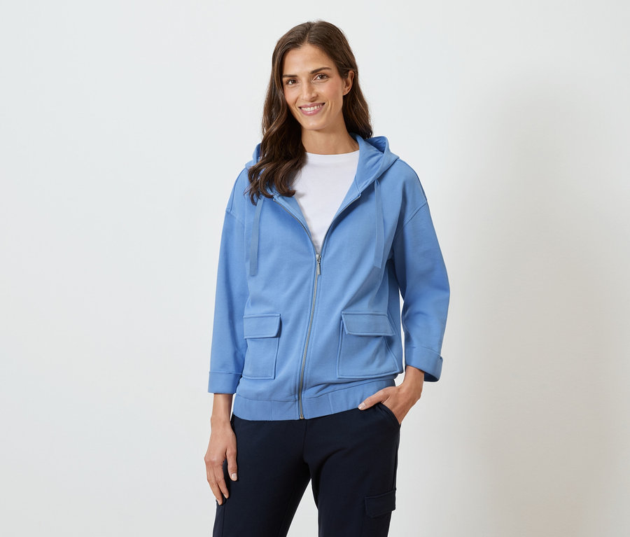 Žena s dlhými hnedými vlasmi sa usmieva v modrej mikine s kapucňou a tmavých nohaviciach.