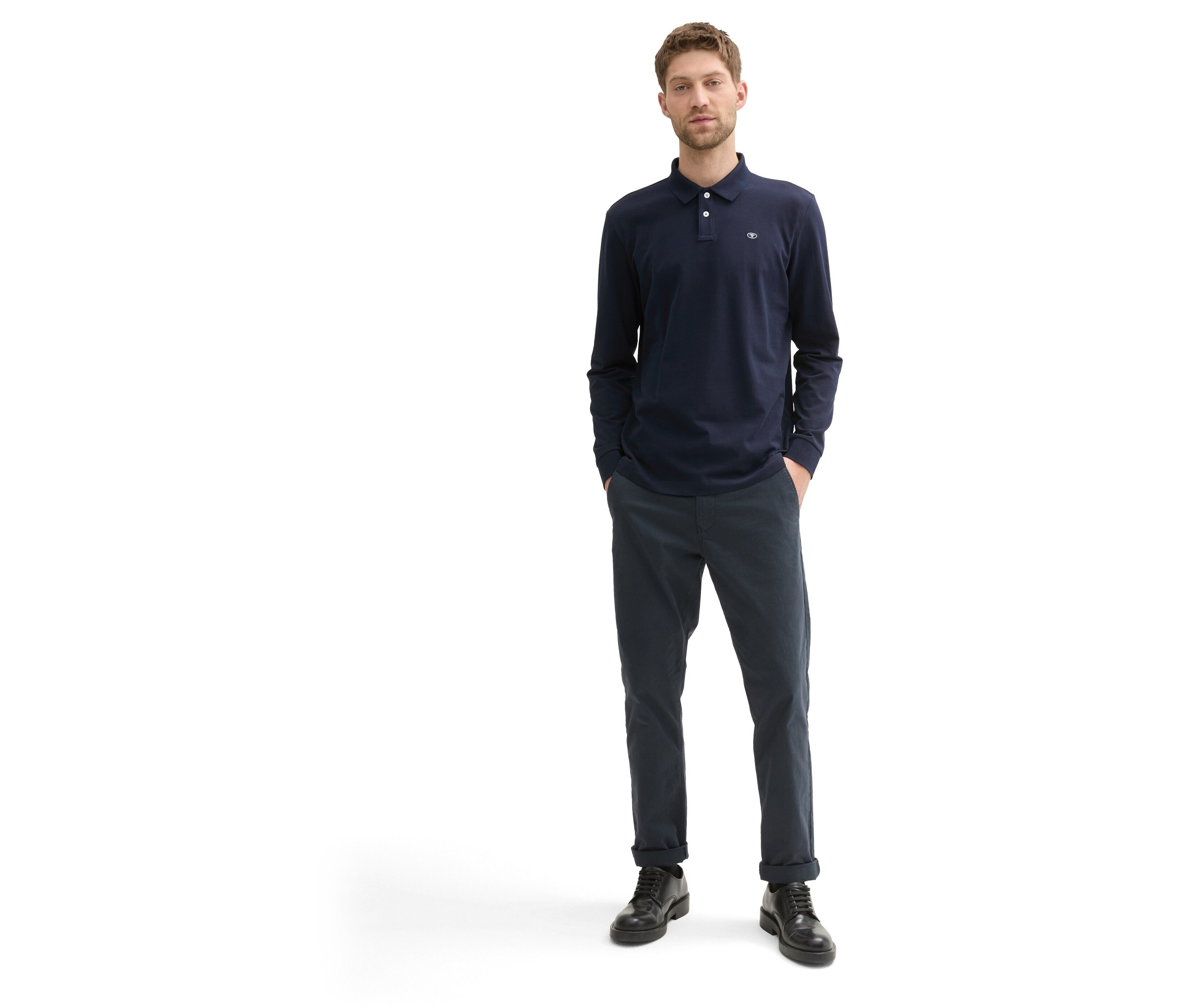 Muž stojí a má na sebe tmavomodré tričko s dlhým rukávom, tmavomodré nohavice a čierne topánky.