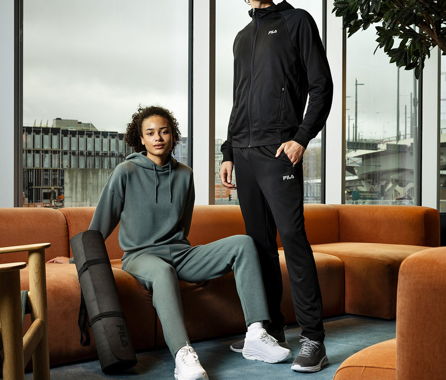 Mladá žena sedí s zrolovanou podložkou na jogu vedľa seba na oranžovej pohovke, vedľa nej stojí mladý muž. Obaja majú na sebe teplákové súpravy a tenisky.