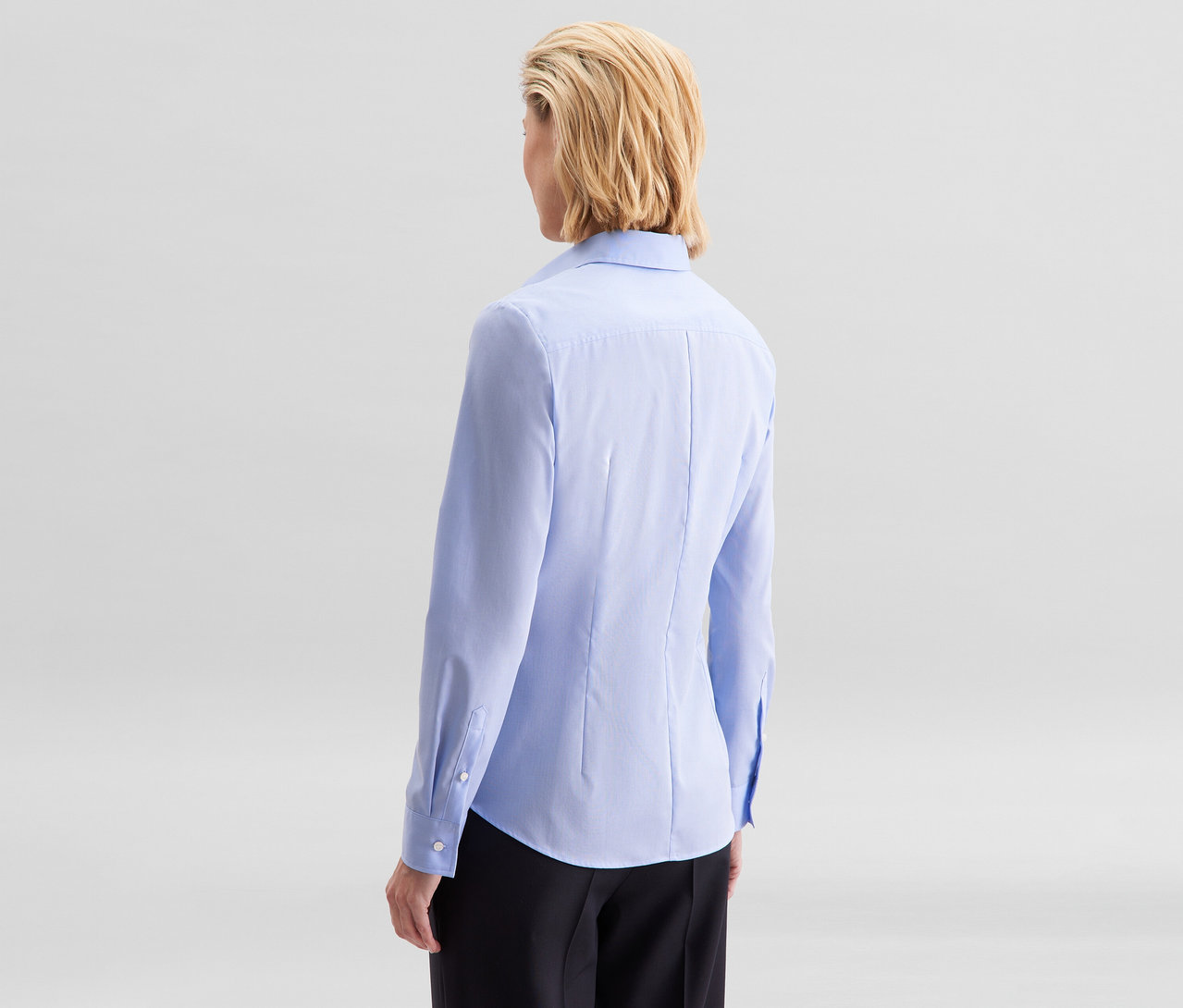 Žena zozadu, s krátkymi blond vlasmi, má na sebe svetlomodrú blúzku Seidensticker Slim Fit non iron a čierne nohavice.