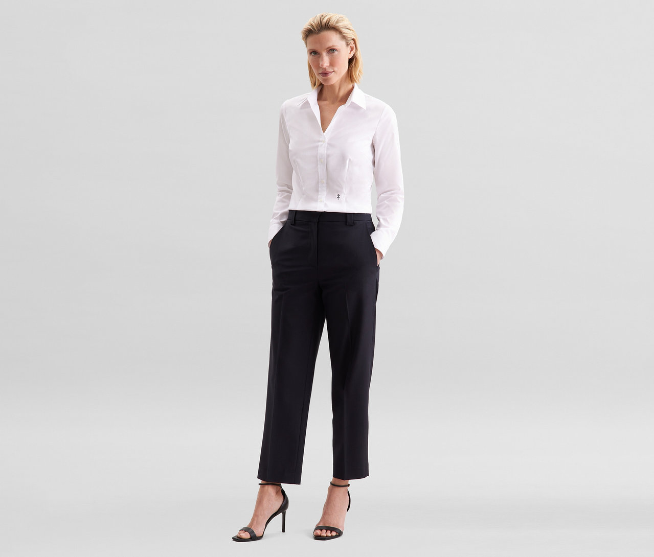 Žena s blond vlasmi stojí s rukami vo vreckách čiernych nohavíc. Má na sebe bielu blúzku Seidensticker Slim Fit non iron a čierne sandále na vysokom opätku.