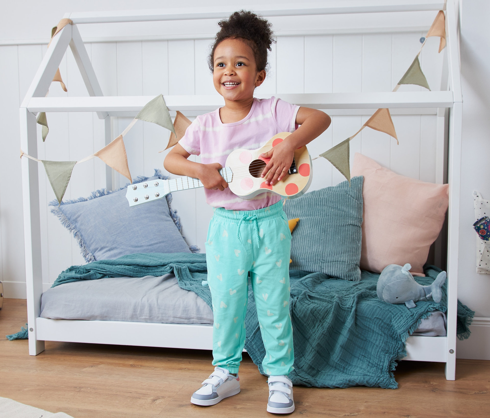 Usmievajúce sa dievčatko hrá na ukulele v detskej izbe. Má na sebe navliekacie nohavice z biobavlny.