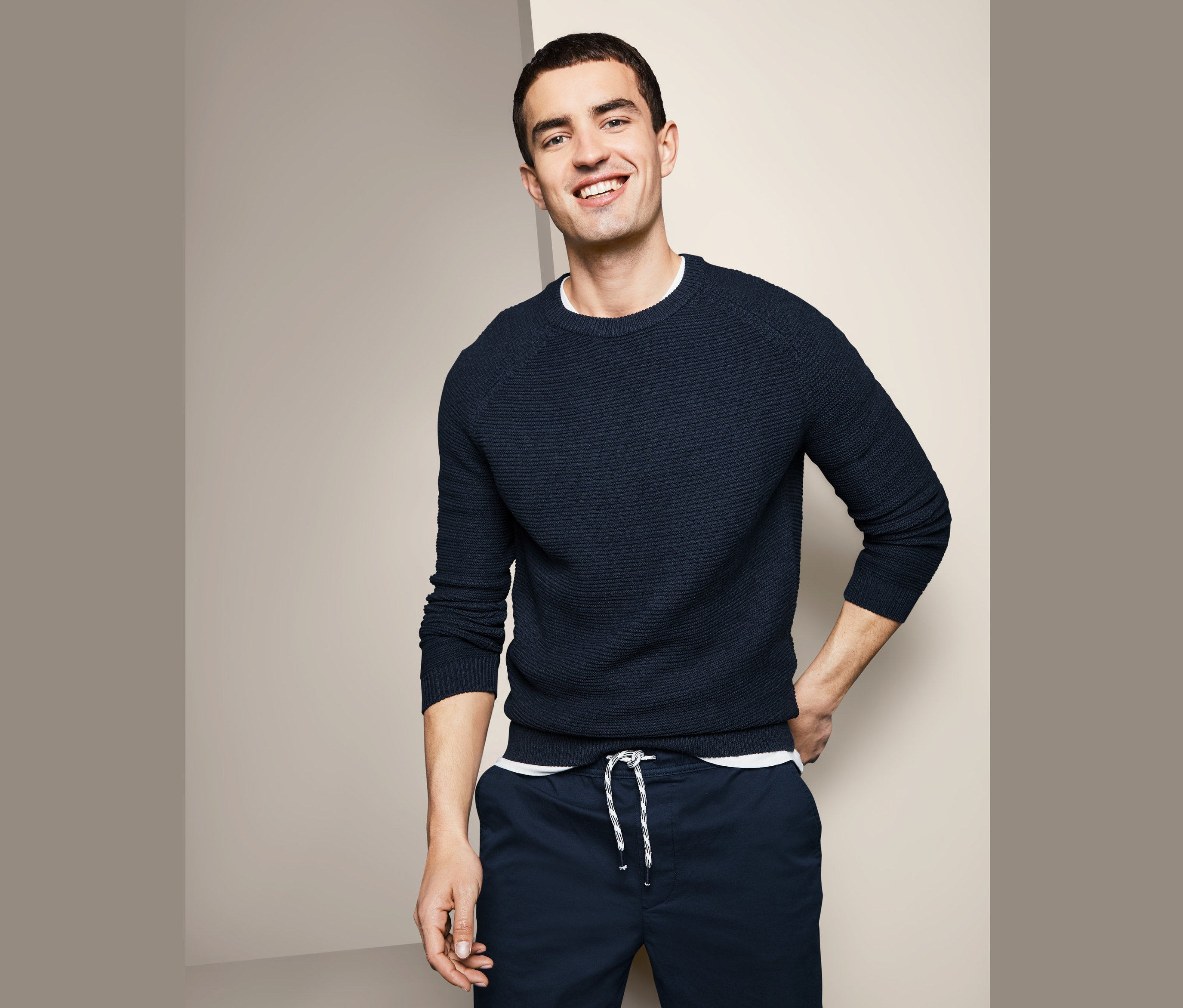 Usmievajúci sa muž pózuje v pulóvri z hrubej pleteniny a tmavomodrých nohaviciach.