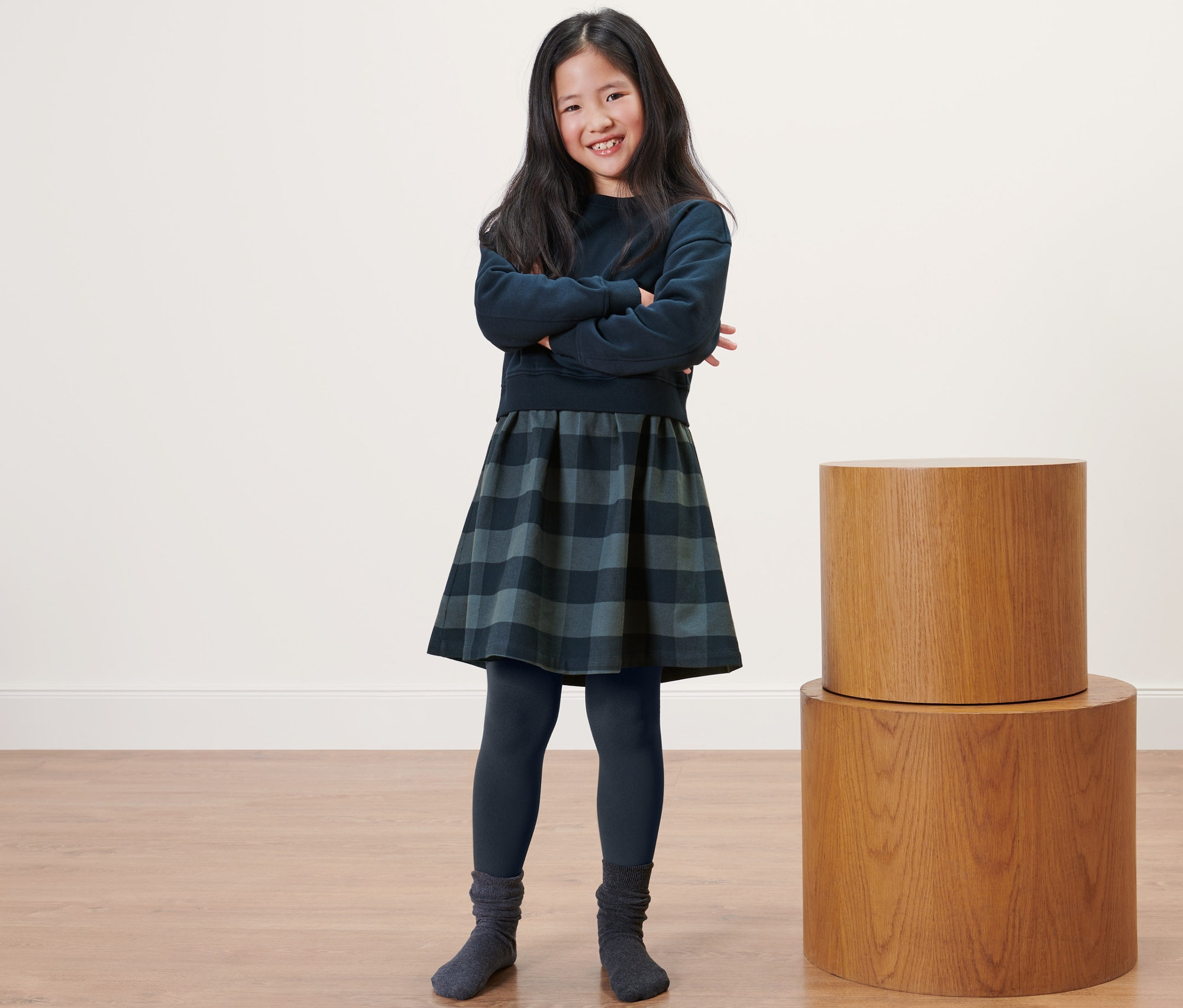 Usmievajúce sa dievča stojí s rukami prekríženými na hrudi a má na sebe detské termo legíny, šaty a sveter.