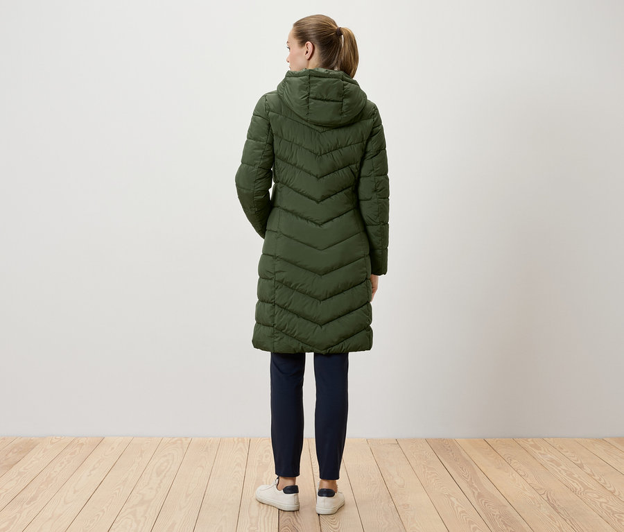 Žena stojí chrbtom k fotoaparátu a má na sebe dlhý zelený prešívaný kabát s kapucňou, tmavomodré nohavice a biele tenisky.
