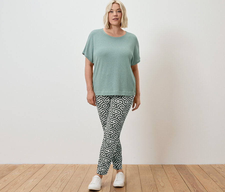 Žena stojí a má na sebe štruktúrované tričko šalviovej farby a zelené džersejové nohavice s potlačou.