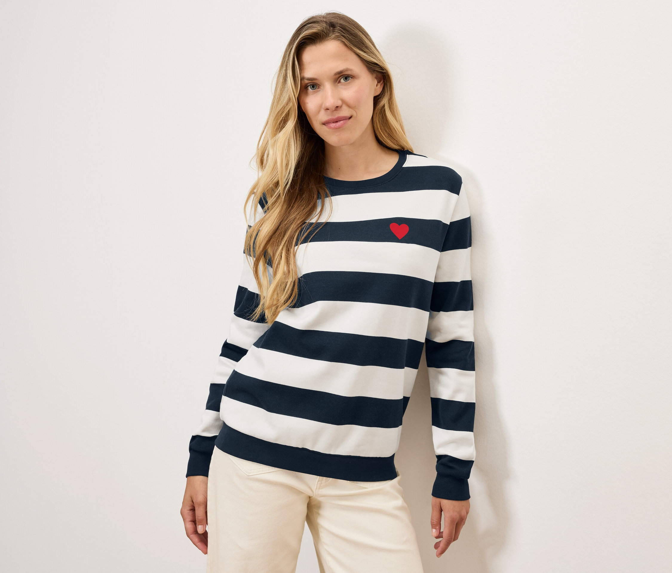 Blondínka pózuje v pruhovanom svetri s červeným srdcom a svetlých nohaviciach.