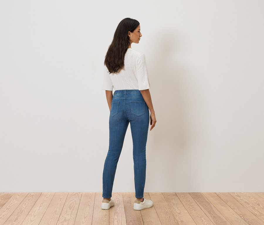 Žena stojí chrbtom k fotoaparátu a má na sebe modré rifle, biely top a biele tenisky.