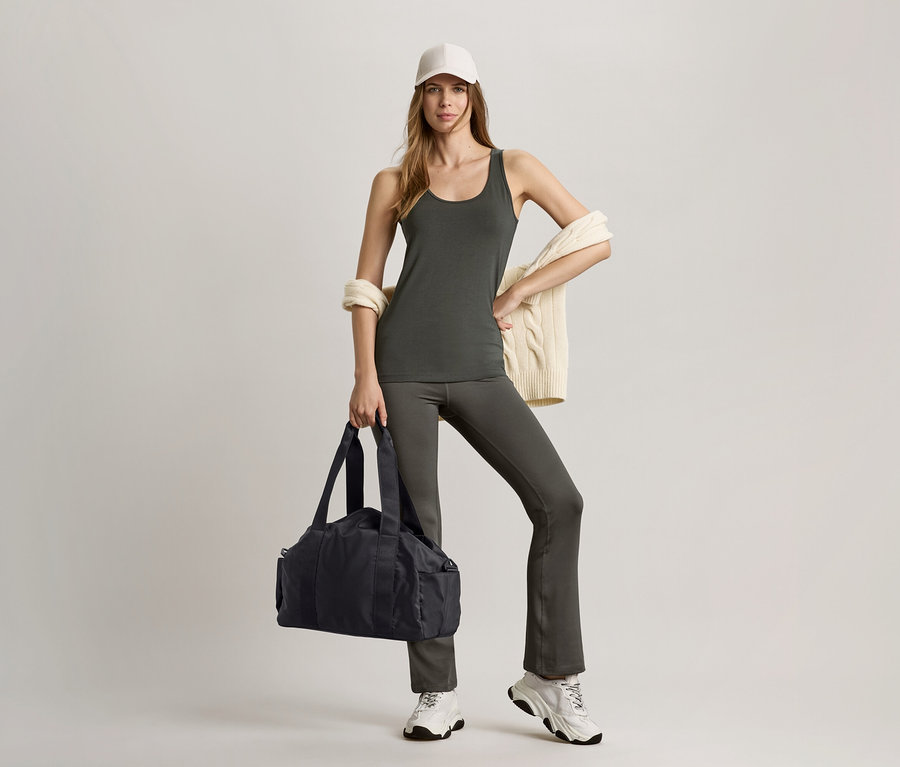 Žena v olivovozelenom tielku a sťahovacích športových nohaviciach drží čiernu športovú tašku.