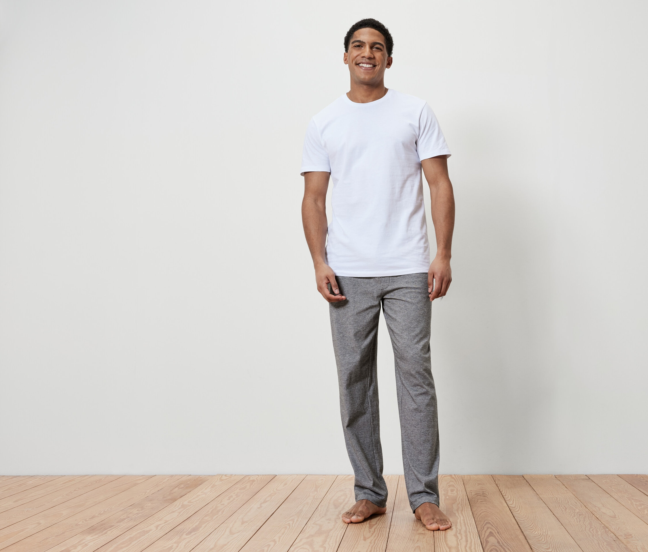 Usmievajúci sa muž stojí naboso na drevenej podlahe. Má na sebe biele tričko a sivé nohavice.