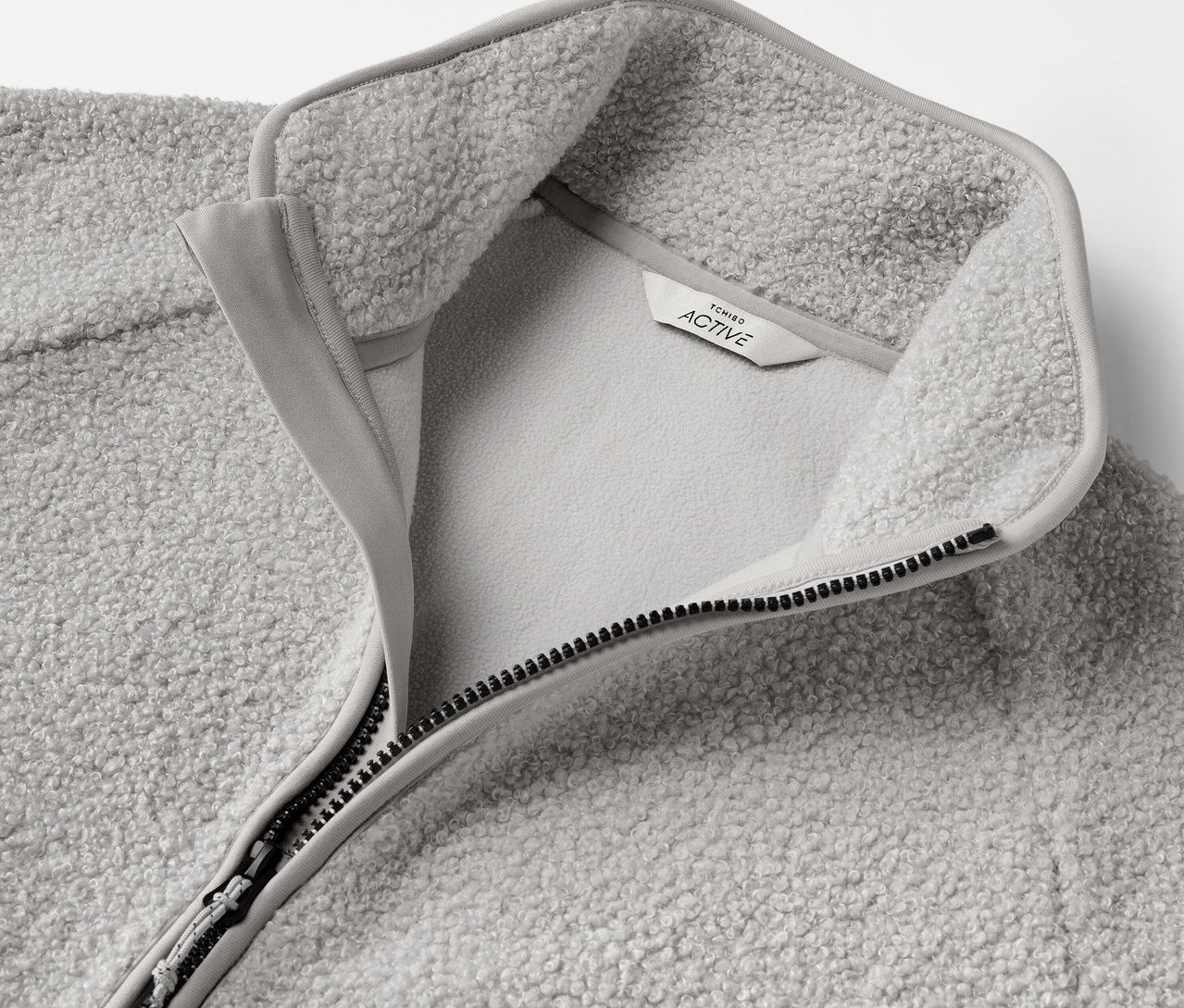 Detail šedej fleecovej mikiny s rozopnutým zipsom a štítkom.