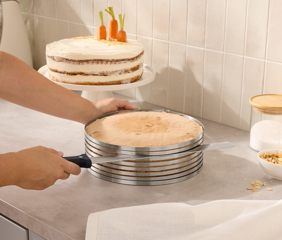 Osoba krája tortu pomocou nastaviteľného krájača na torty. V pozadí je torta s mrkvou a poháre s viečkami.