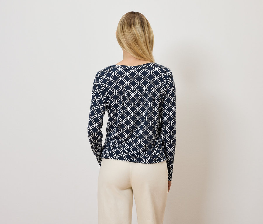 Žena chrbtom k fotoaparátu má na sebe tmavomodré tričko s dlhým rukávom a geometrickým vzorom a béžové nohavice.