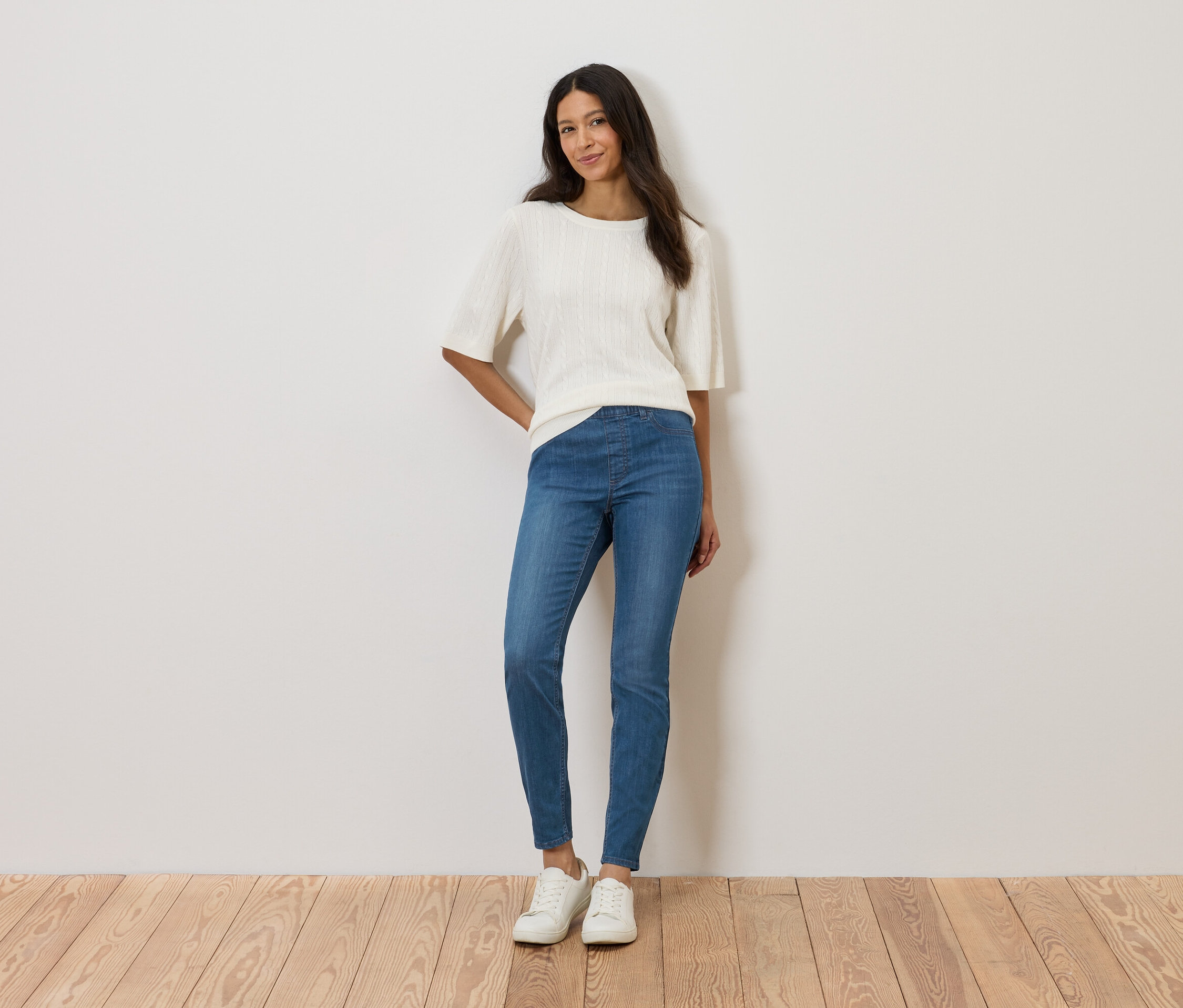 Žena stojí pred stenou a má na sebe biely top, modré džínsy a biele topánky.