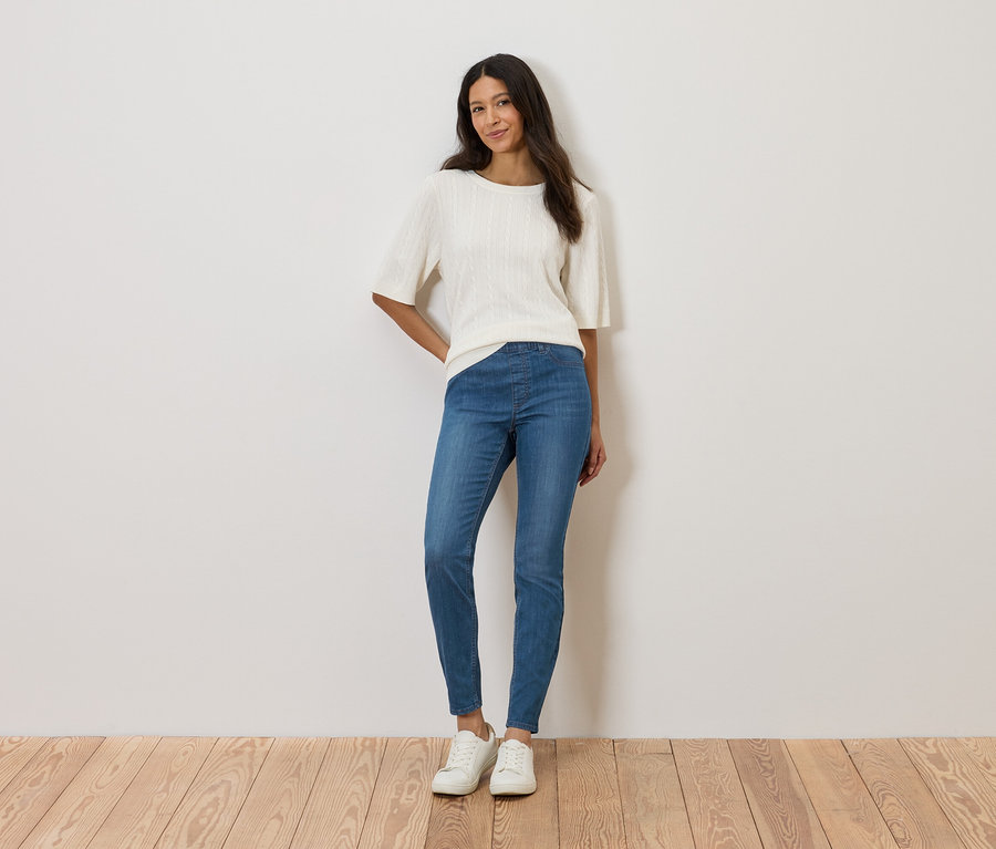 Žena stojí pred stenou a má na sebe biely top, modré džínsy a biele topánky.