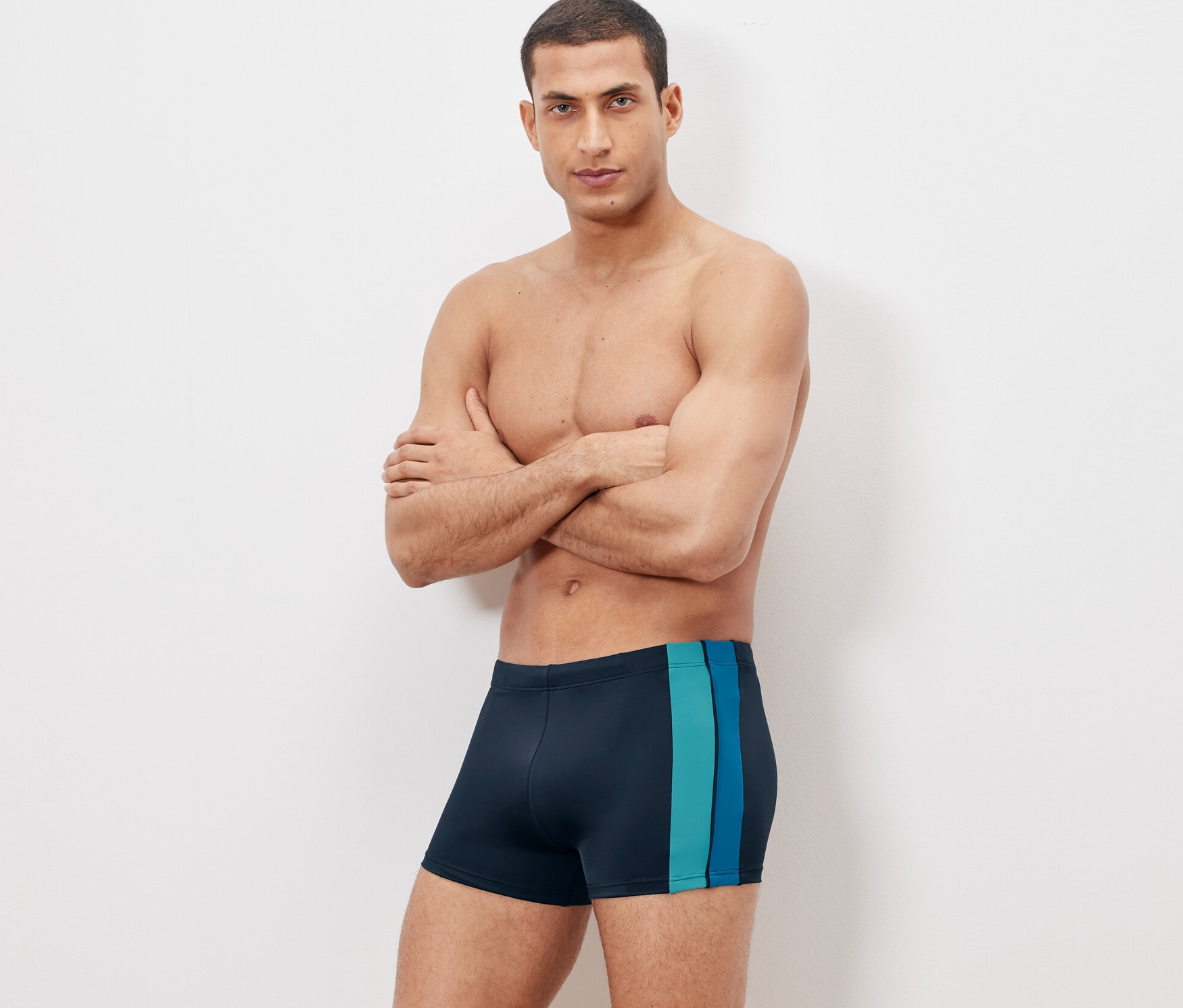 Muž pózuje so skríženými rukami v plávacích šortkách.