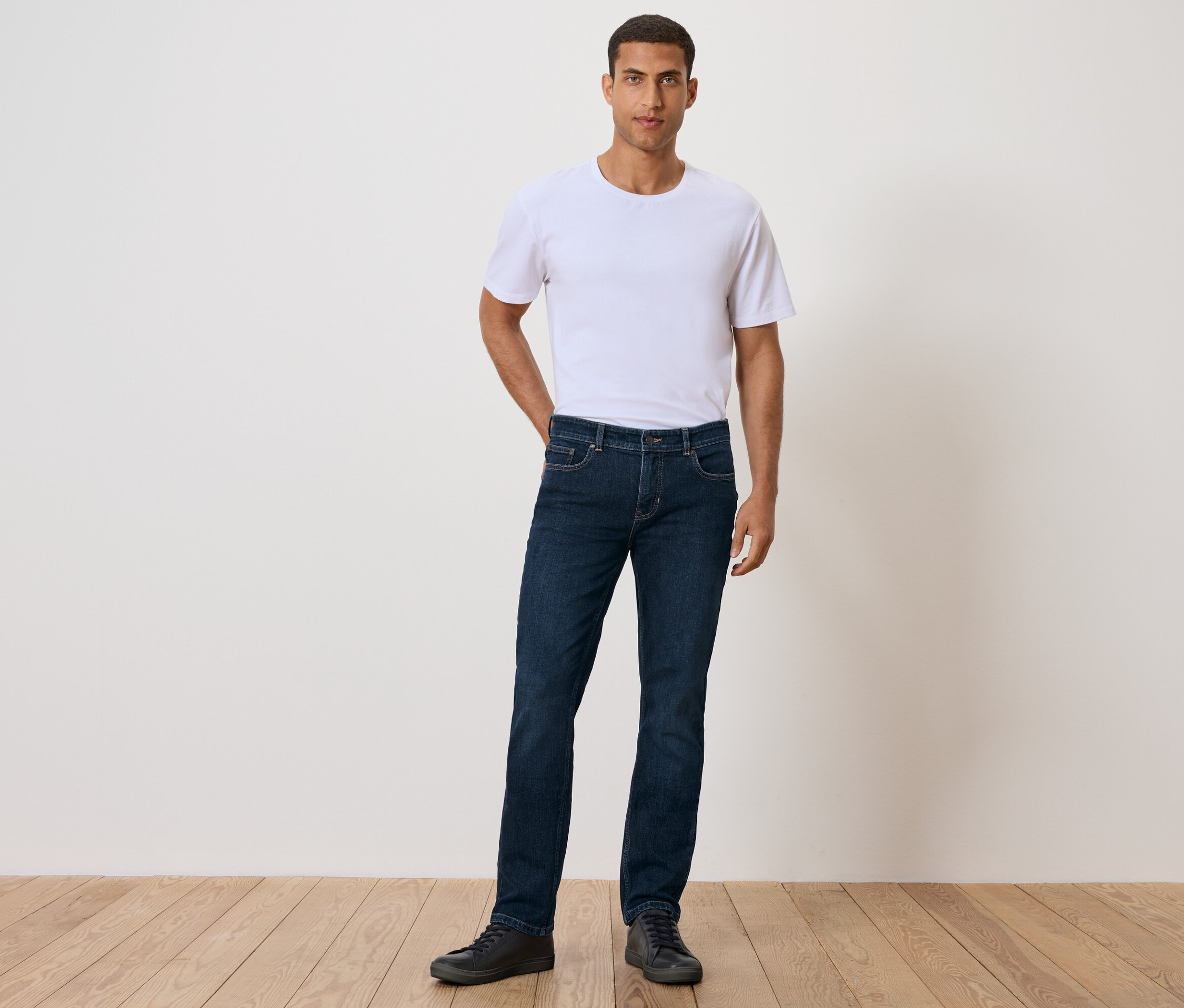 Muž stojí s rukou vo vrecku, má na sebe biele tričko, džínsy a čierne topánky.