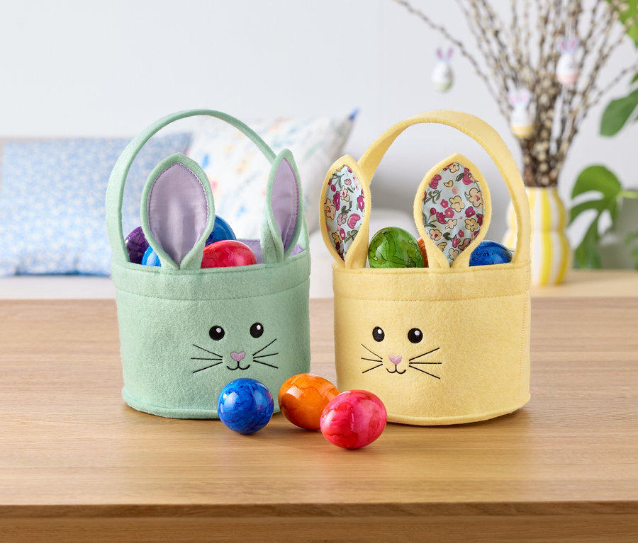 Dva veľkonočné košíky v tvare zajačika, jeden mätovo zelený a jeden žltý, plné farebných vajec, stoja na stole.