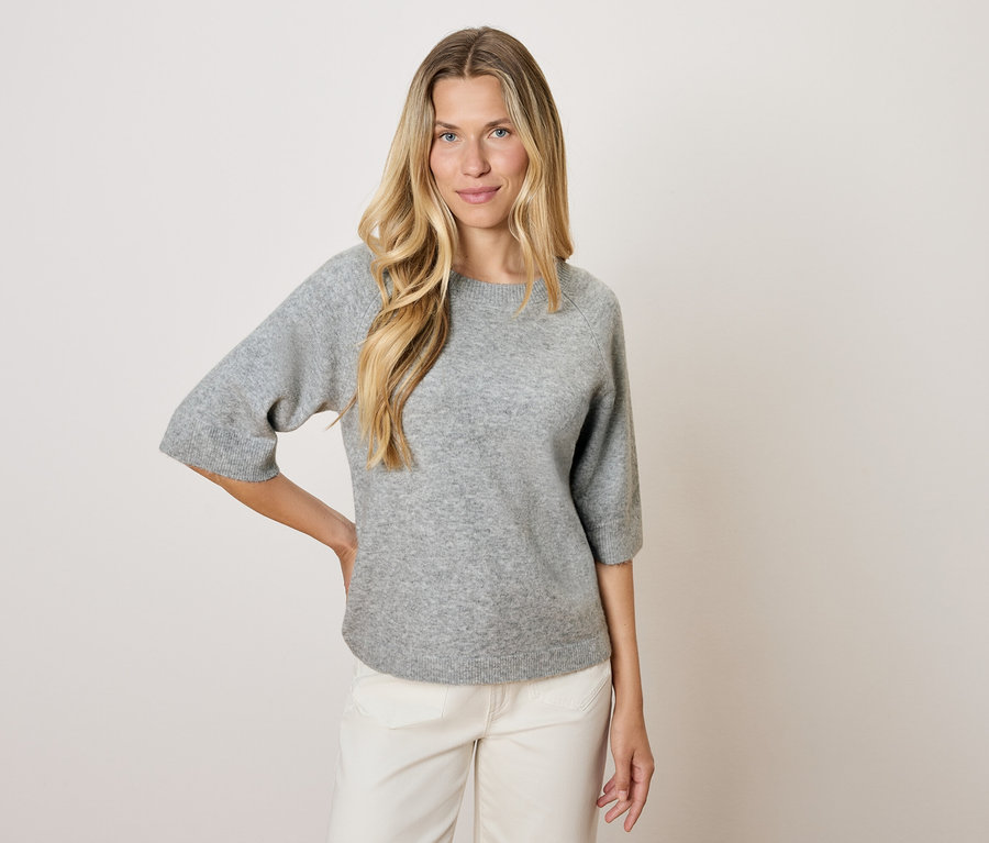 Blondínka v sivom svetri s trojštvrťovými rukávmi a bielych nohaviciach pózuje na svetlom pozadí.