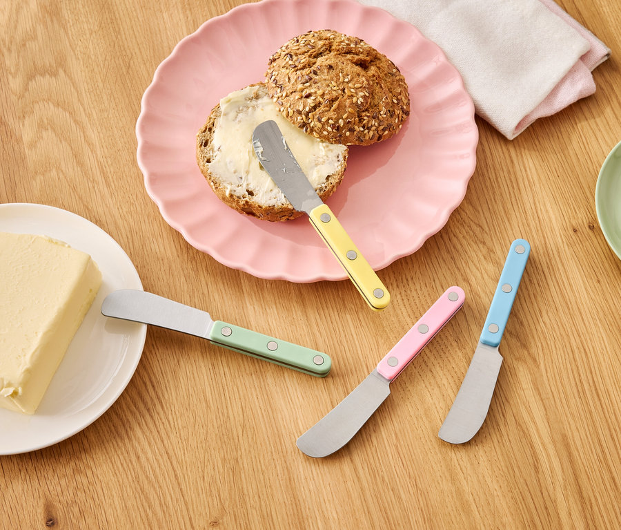 Rožok s maslom na ružovom tanieri vedľa kocky masla a farebných nožov na drevenom stole.