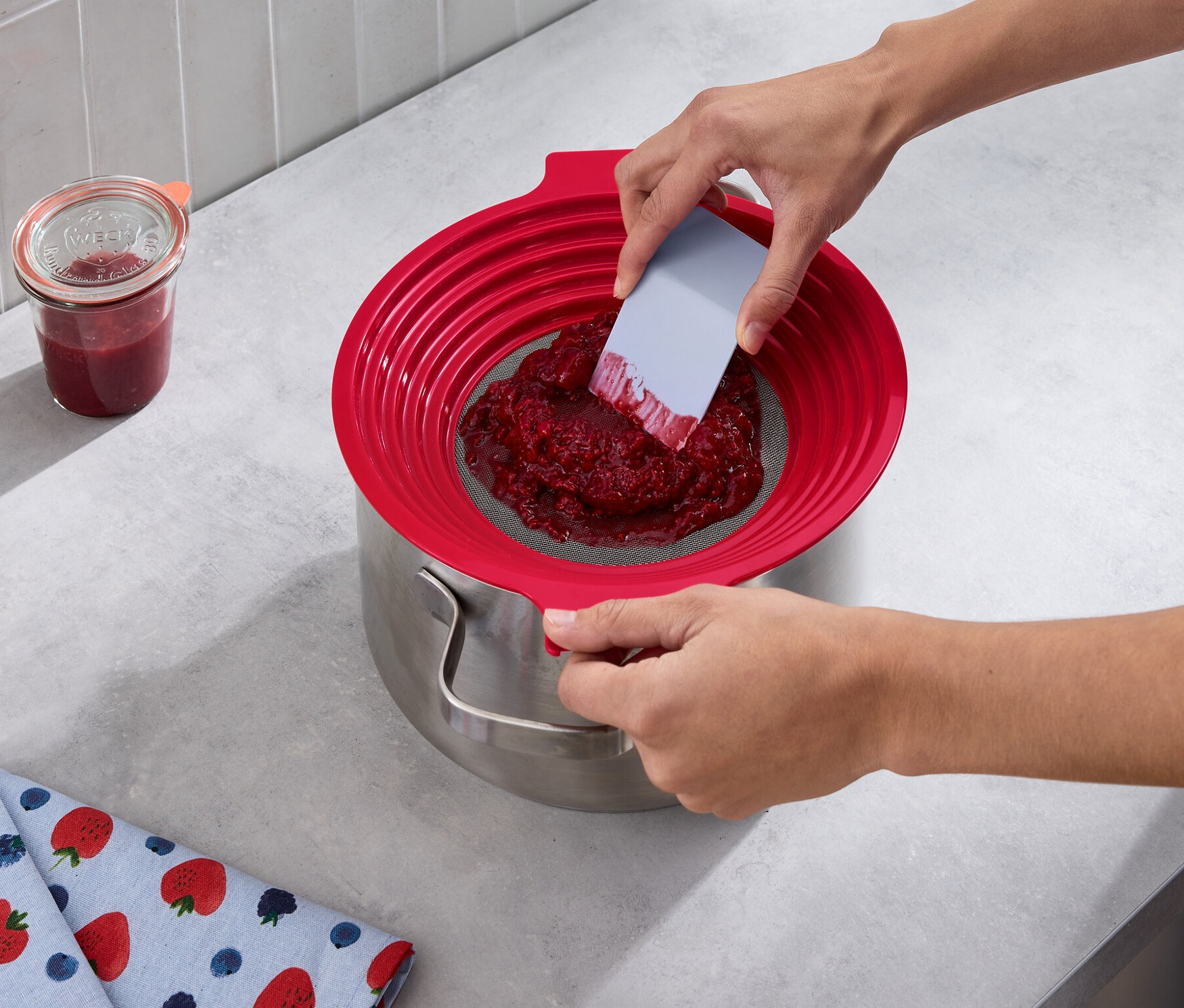 Osoba pasíruje červené ovocie cez 2 v 1: sito na preosievanie múky a pasírovanie do hrnca. Vedľa stojí pohár džemu a leží utierka s potlačou jahôd.