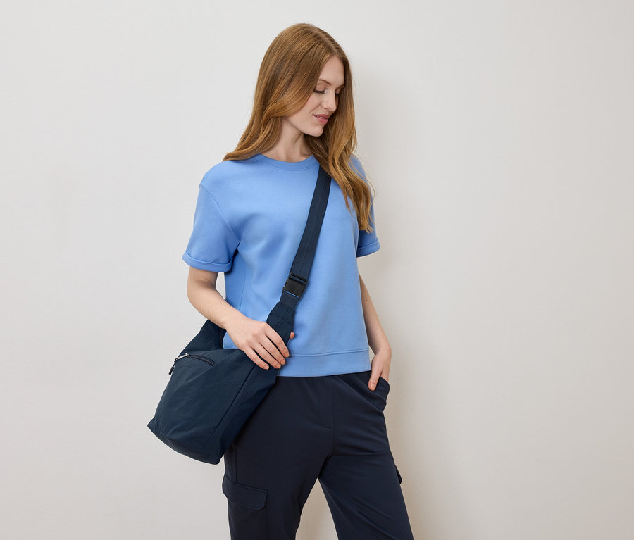 Žena so svetlohnedými vlasmi, svetlomodrým tričkom a námorníckou modrou taškou cez rameno na krémovom pozadí.