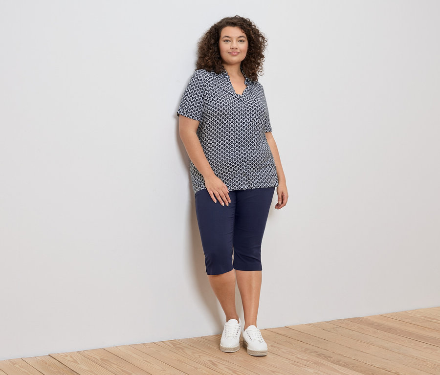 Žena s kučeravými vlasmi sa opiera o stenu a má na sebe modro-bielu blúzku so vzorom, modré capri nohavice a biele tenisky.
