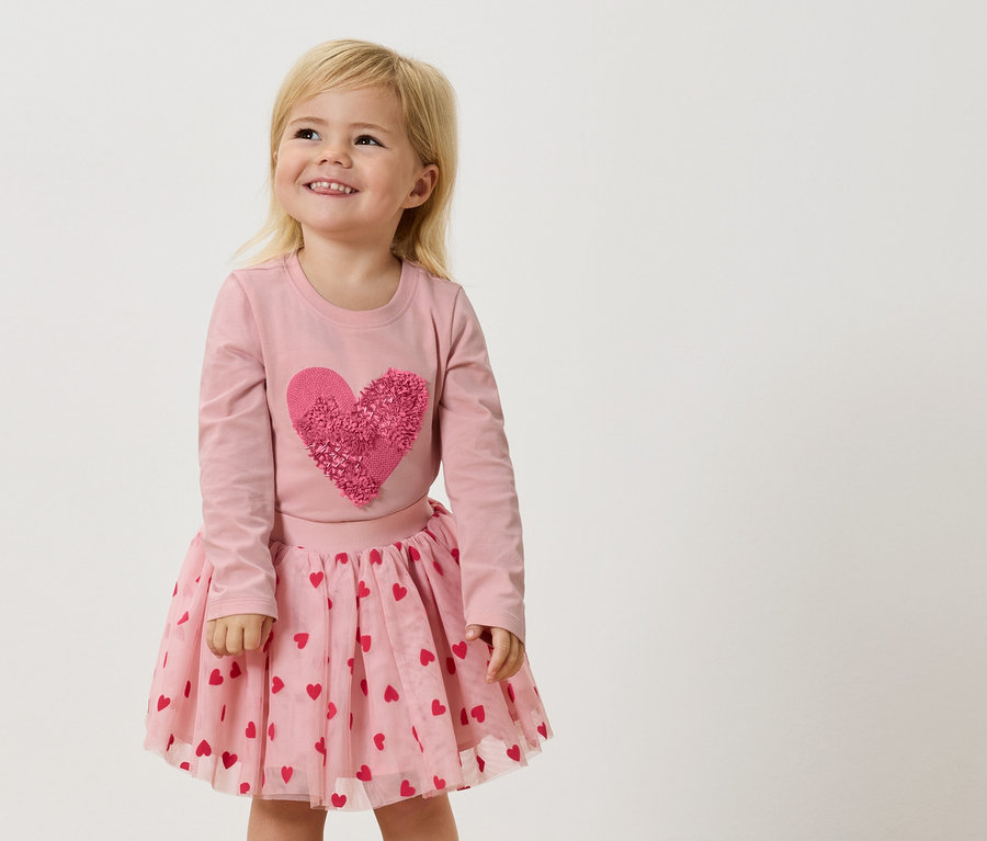 Malé blond dievča má na sebe ružové tričko s dlhým rukávom s veľkým ružovým srdcom a ružovú tylovú sukňu s červenými srdiečkami.