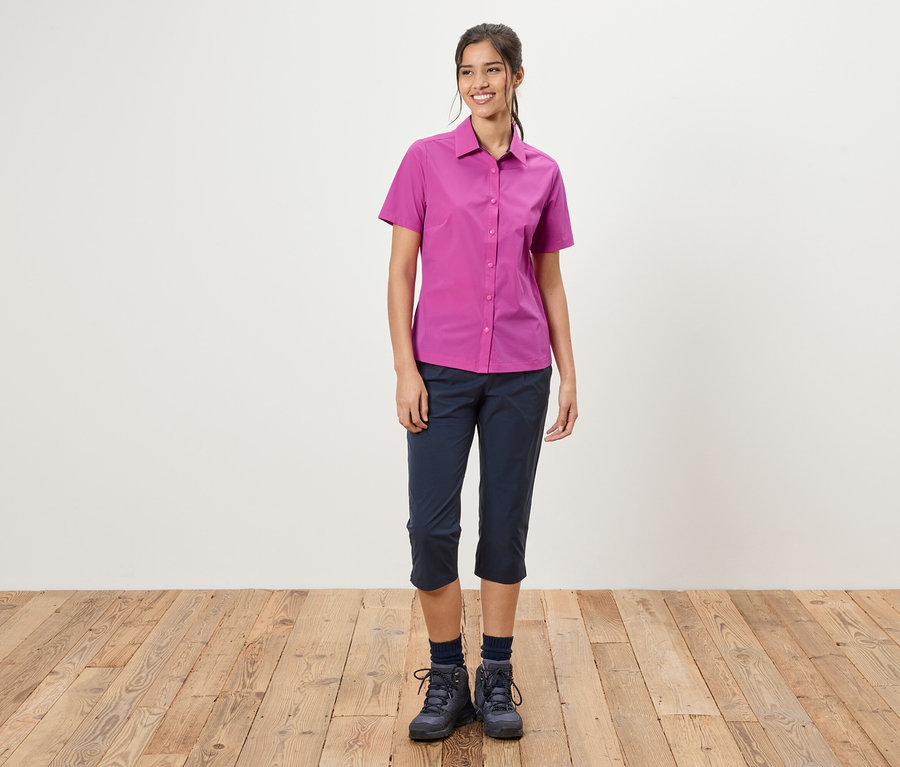 Žena stojí pred bielou stenou. Má na sebe ružovú košeľu, modré trojštvrťové nohavice a turistické topánky.