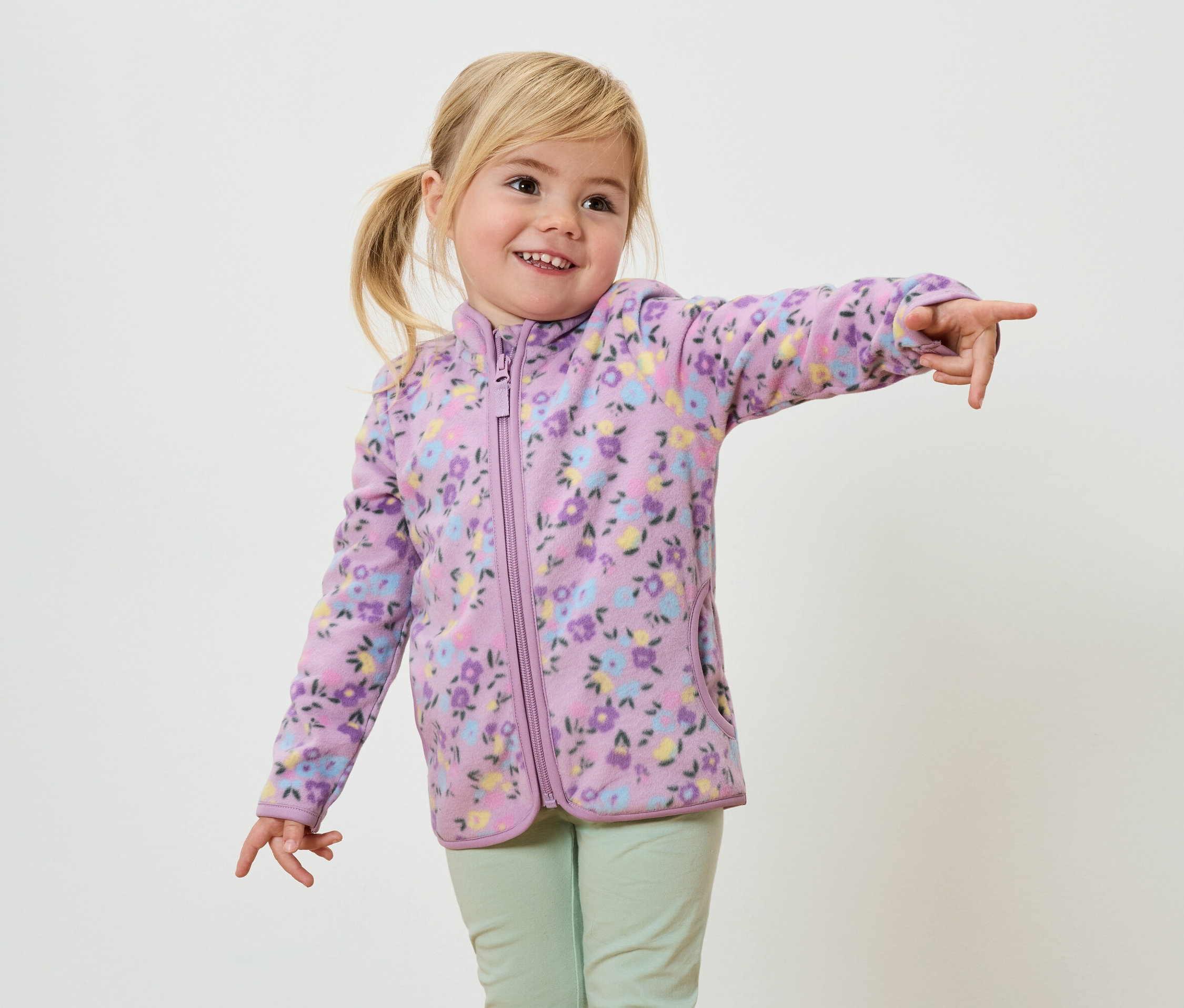 Usmievavé dievča s blond vrkočmi, oblečené v fialovej fleecovej bunde s kvetinovou potlačou a svetlozelených nohaviciach, ukazuje prstom.