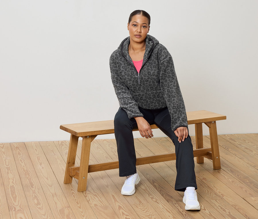 Žena sedí na drevenej lavici a má na sebe sivú flísovú mikinu s leopardím vzorom, ružový top, tmavé nohavice a biele tenisky.