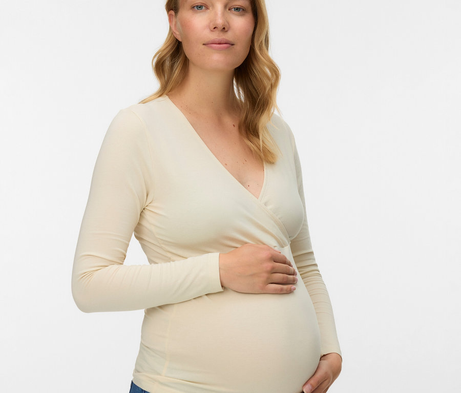 Tehotná žena s blond, vlnitými vlasmi má na sebe blúzku s dlhým rukávom a džínsy.