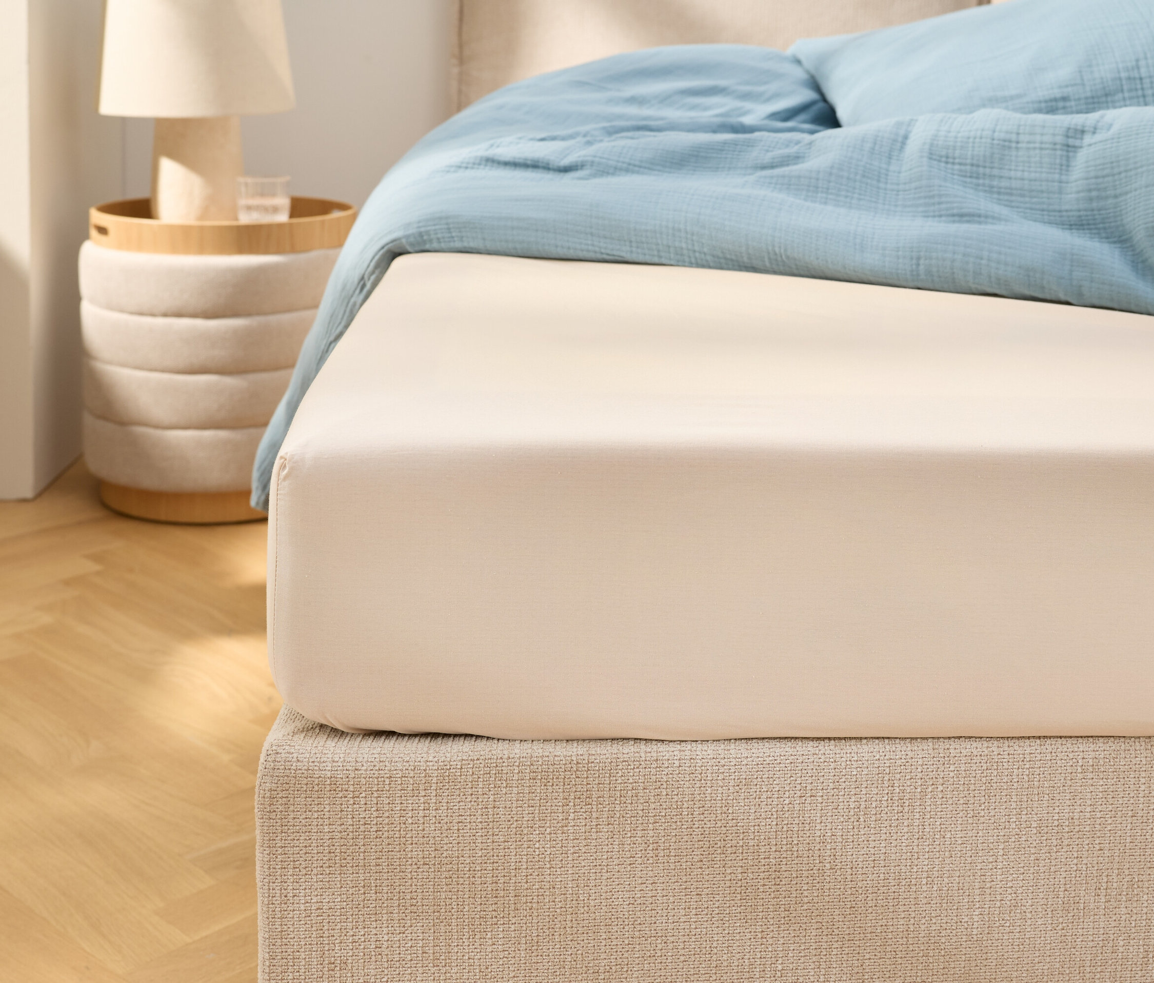 Detailný záber béžovej napínacej plachty na posteli so svetlomodrou posteľnou bielizňou. Vedľa stojí nočný stolík s lampou.