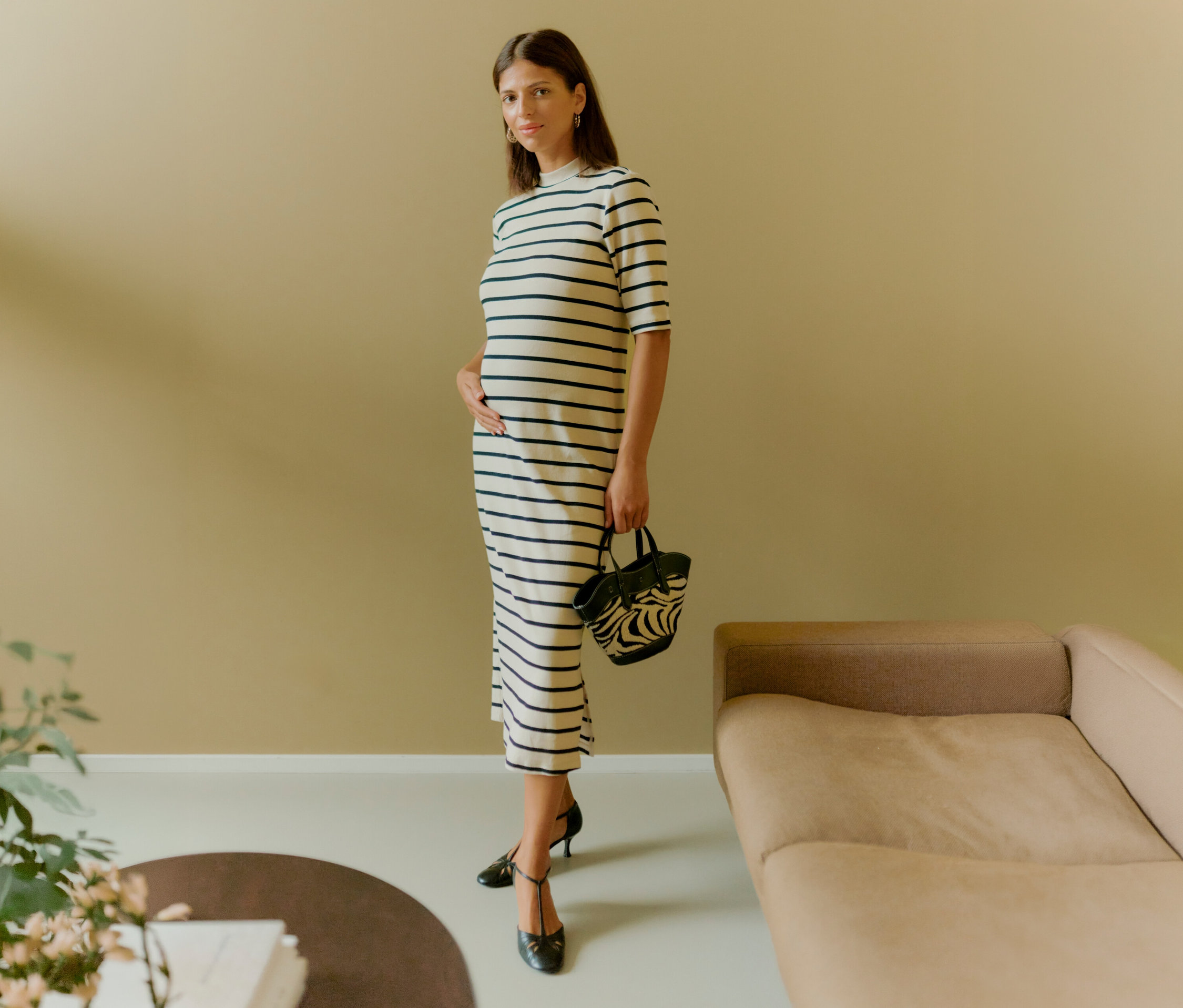 Žena stojí a drží zebra kabelku. Má na sebe pruhované šaty a čierne topánky.