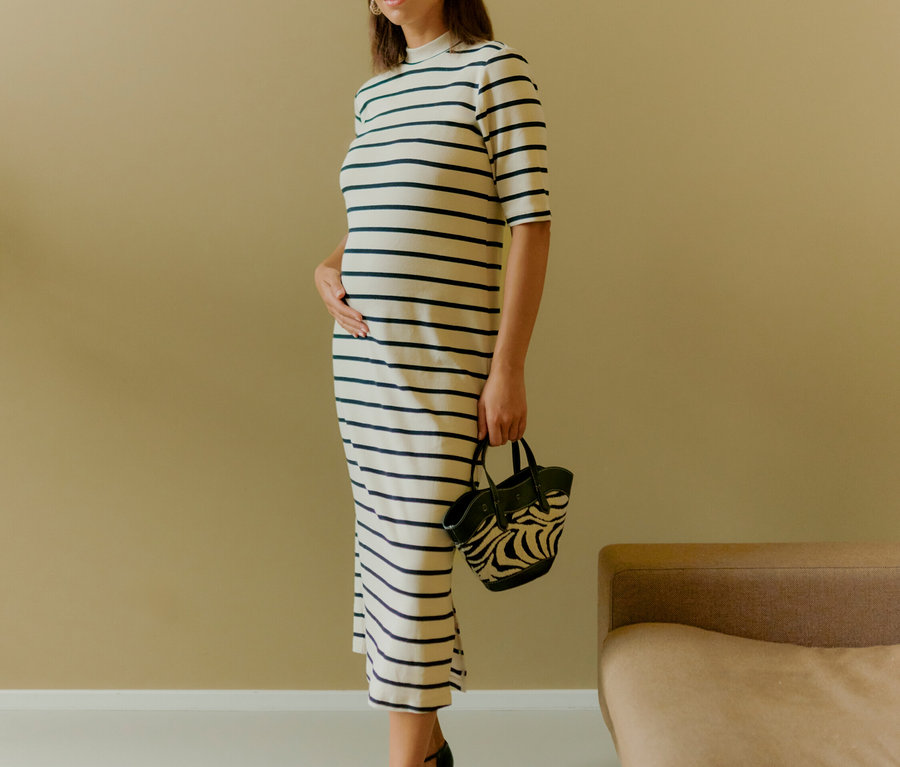 Žena stojí a drží zebra kabelku. Má na sebe pruhované šaty a čierne topánky.