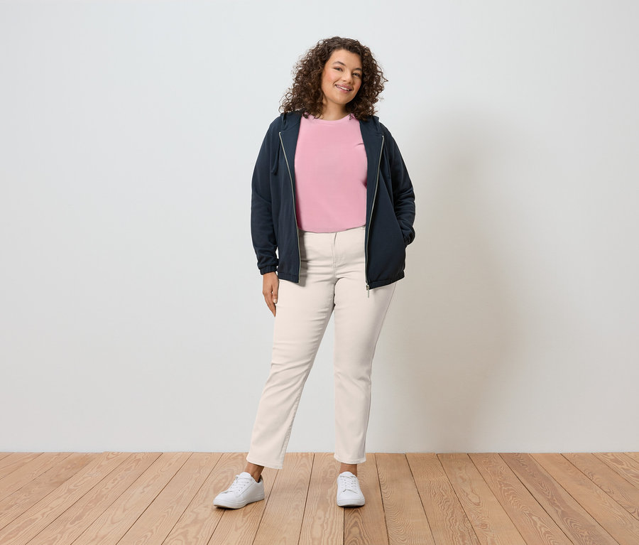 Žena s kučeravými vlasmi stojí pred stenou a má na sebe tmavomodrú bundu s kapucňou, ružové tričko, krémové nohavice a biele tenisky.